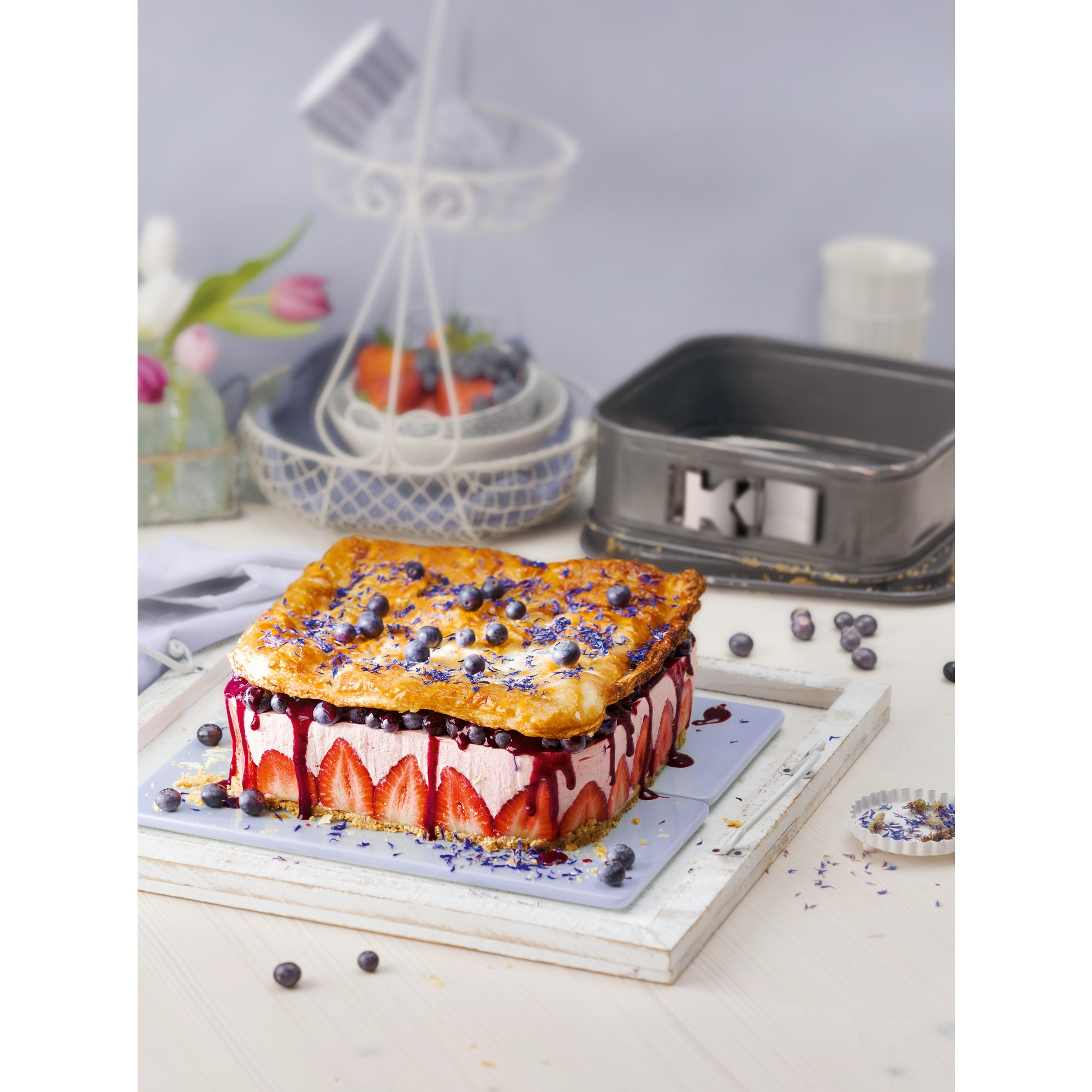 Ciasto z borówkami i truskawkami. Ciasto na jasnoniebieskiej tacy i białym stole.