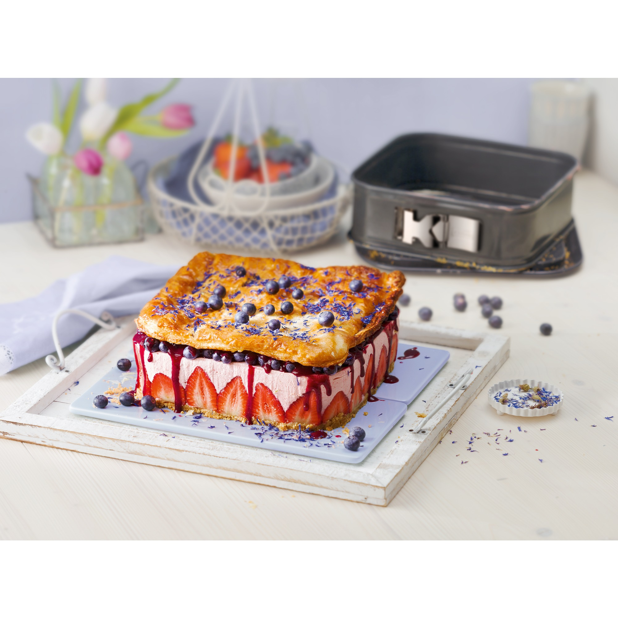 Ciasto z truskawkami, śmietaną i borówkami. Leży na białej tacy. Tło z kwiatami i patelnią.