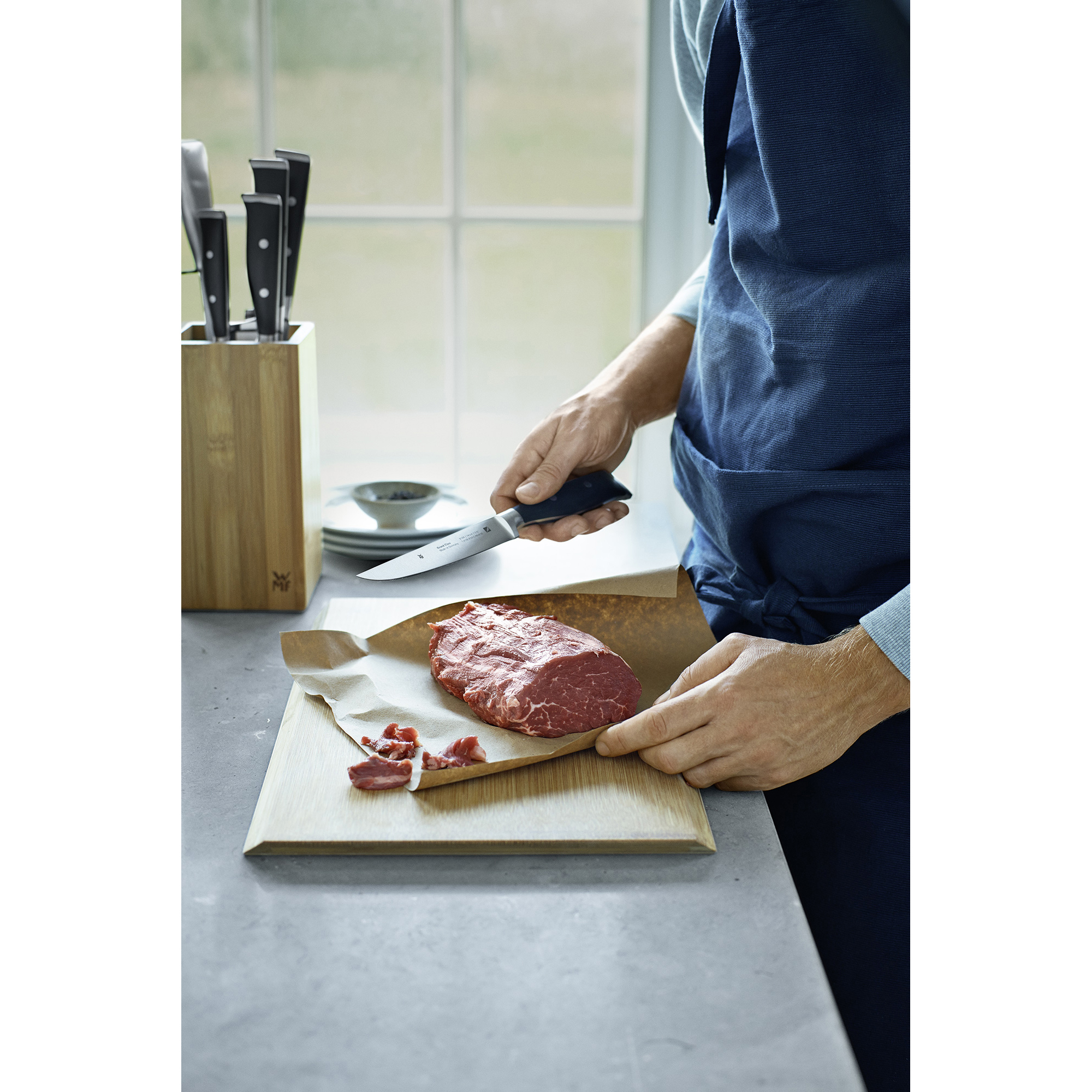 Eine Person schneidet ein rohes Steak auf einem Holzschneidebrett neben einem Messerblock.