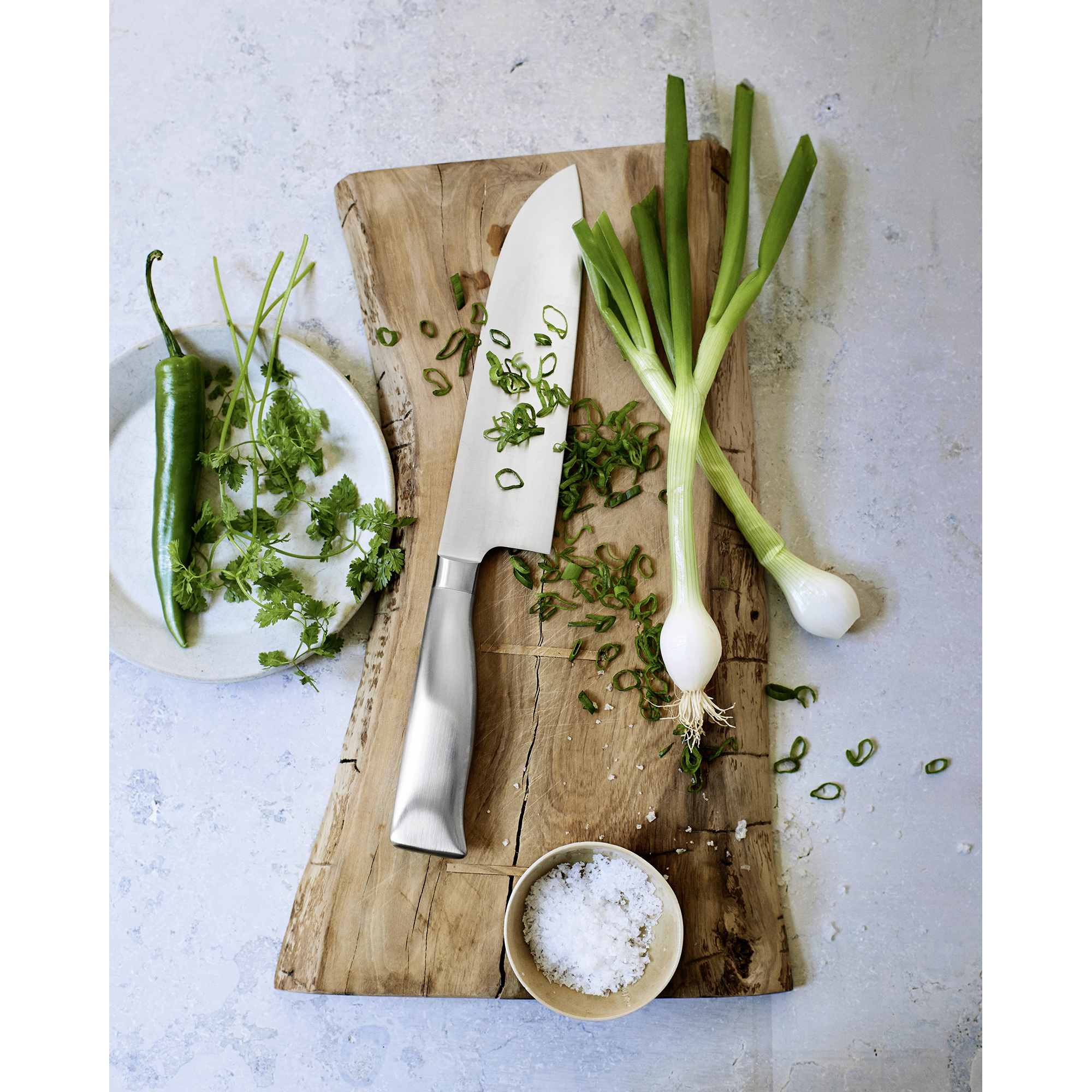 Nóż na drewnianej desce z posiekaną zieloną cebulą, zielonym chili, solą i kolendrą na talerzu.