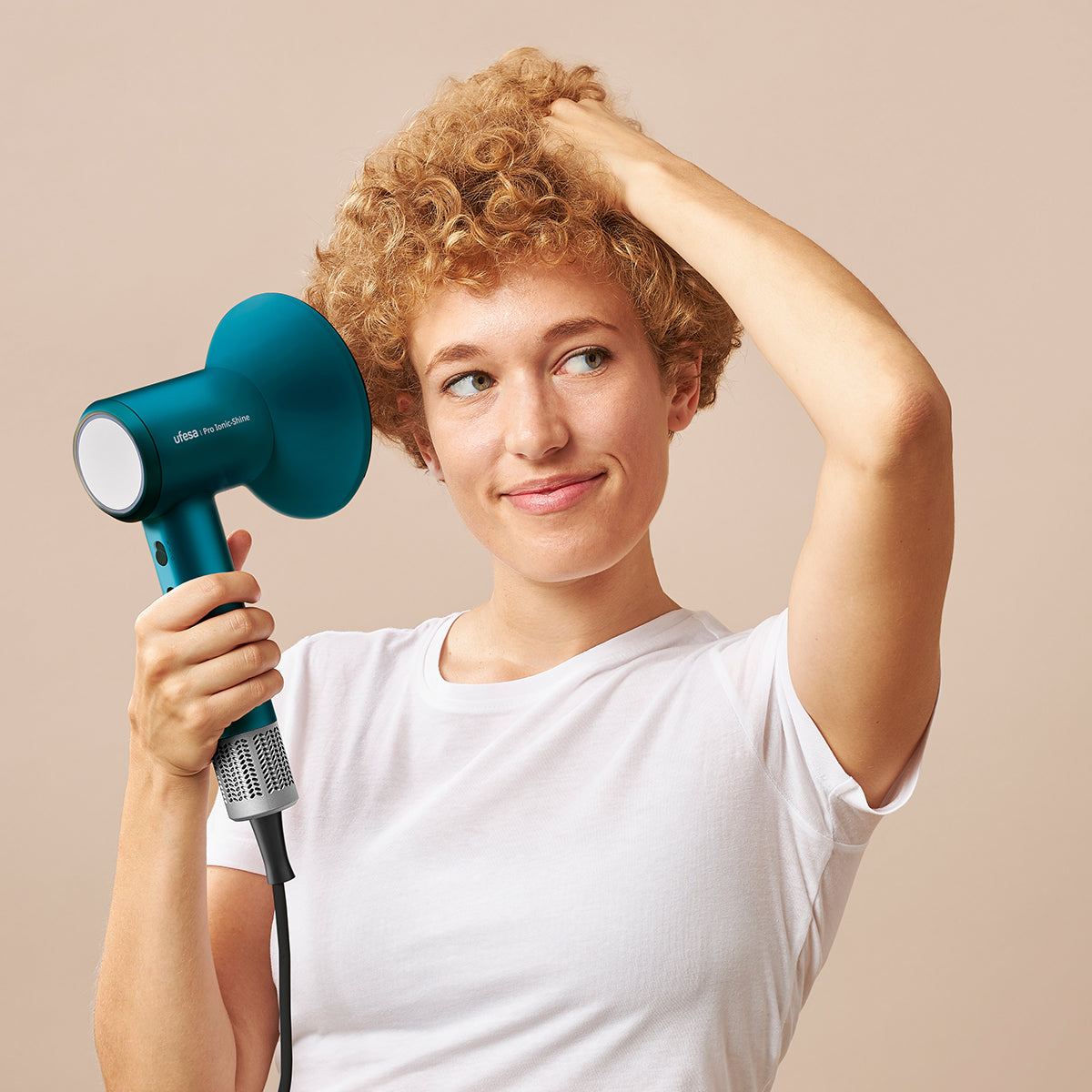 Una mujer con cabello rizado usando un secador turquesa, mirando hacia un lado.
