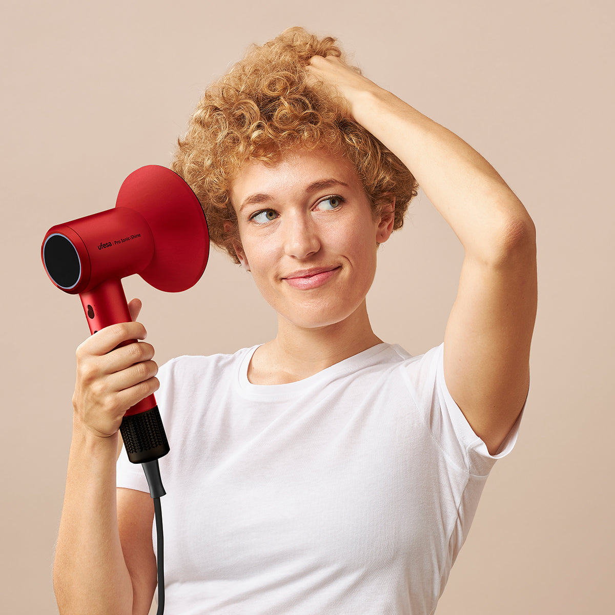Kobieta używa czerwonej suszarki do włosów, trzyma włosy i uśmiecha się w bok.