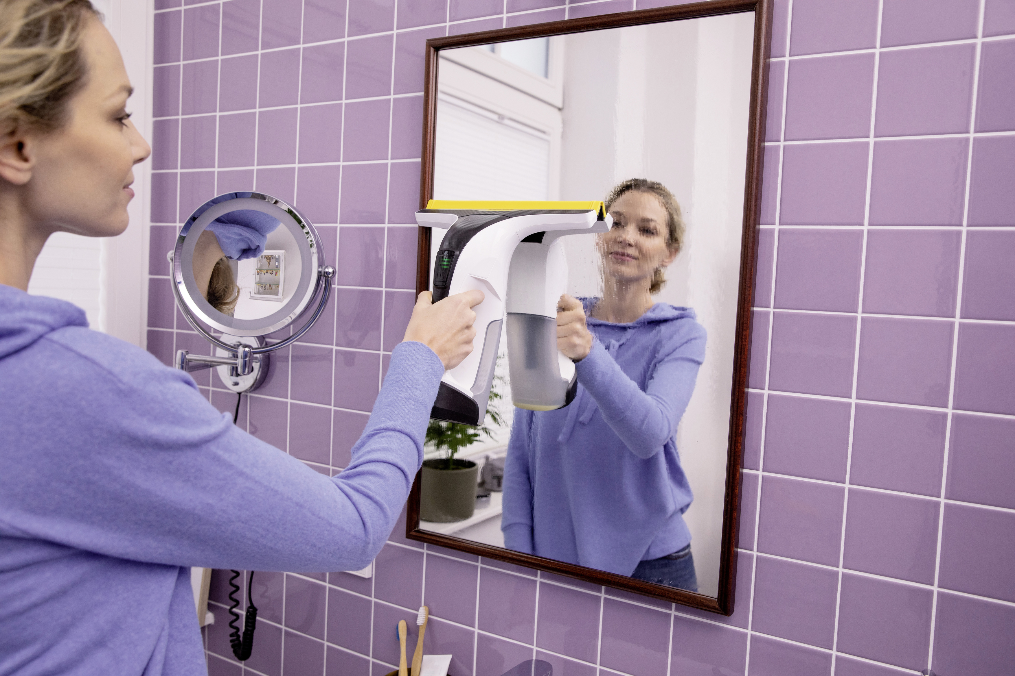 Kobieta czyści lustro za pomocą myjki do okien. Lustro znajduje się w fioletowej łazience w kafelkach.