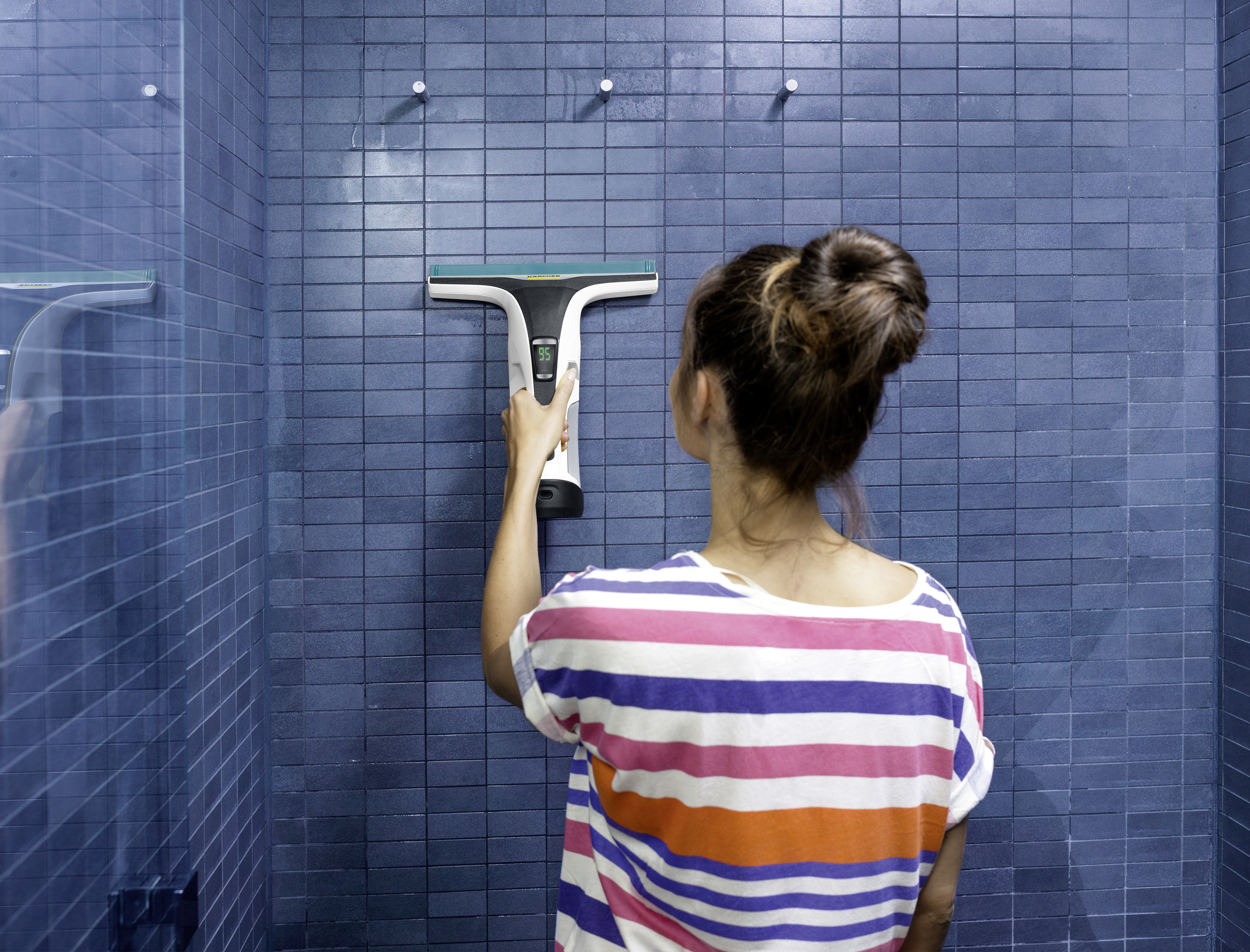 Vrouw maakt blauwe betegelde douche schoon met een wisser. Ze draagt een gestreept shirt.