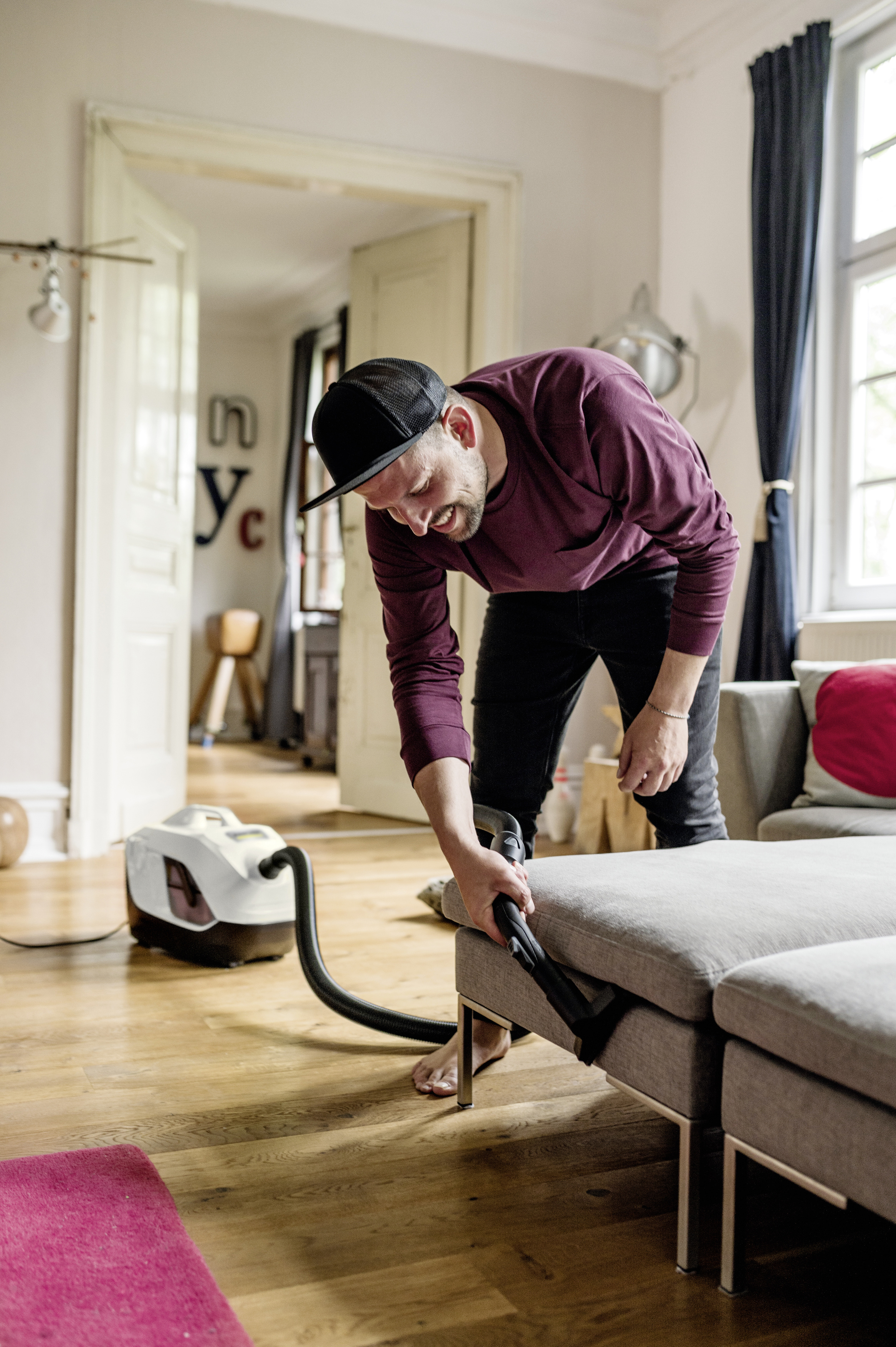 Mężczyzna w czapce, odkurzający sofę; ma na sobie bordową koszulę, w nowoczesnym salonie.