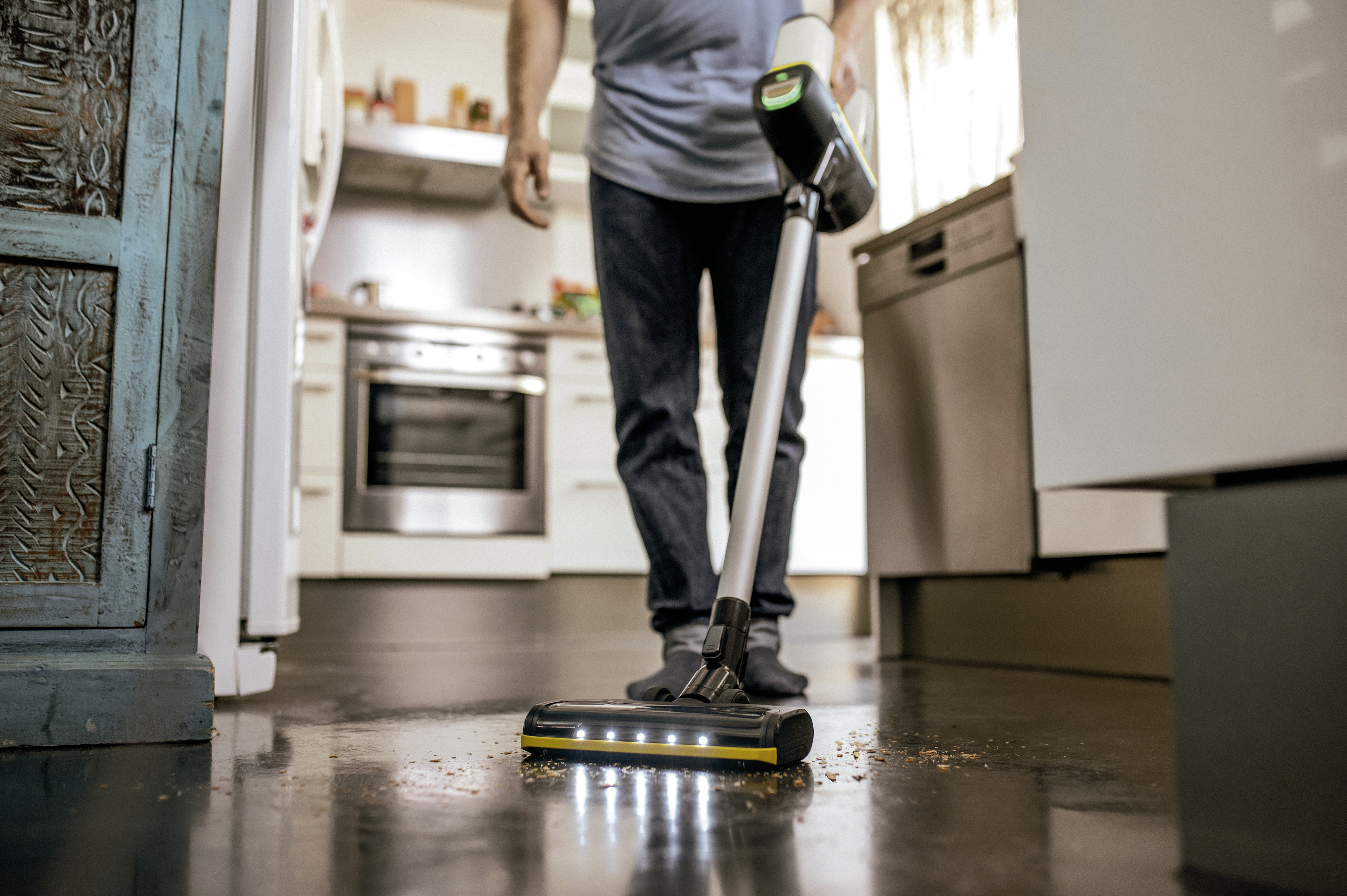 A person vacuuming a dirty floor in a kitchen with a cordless vacuum cleaner.