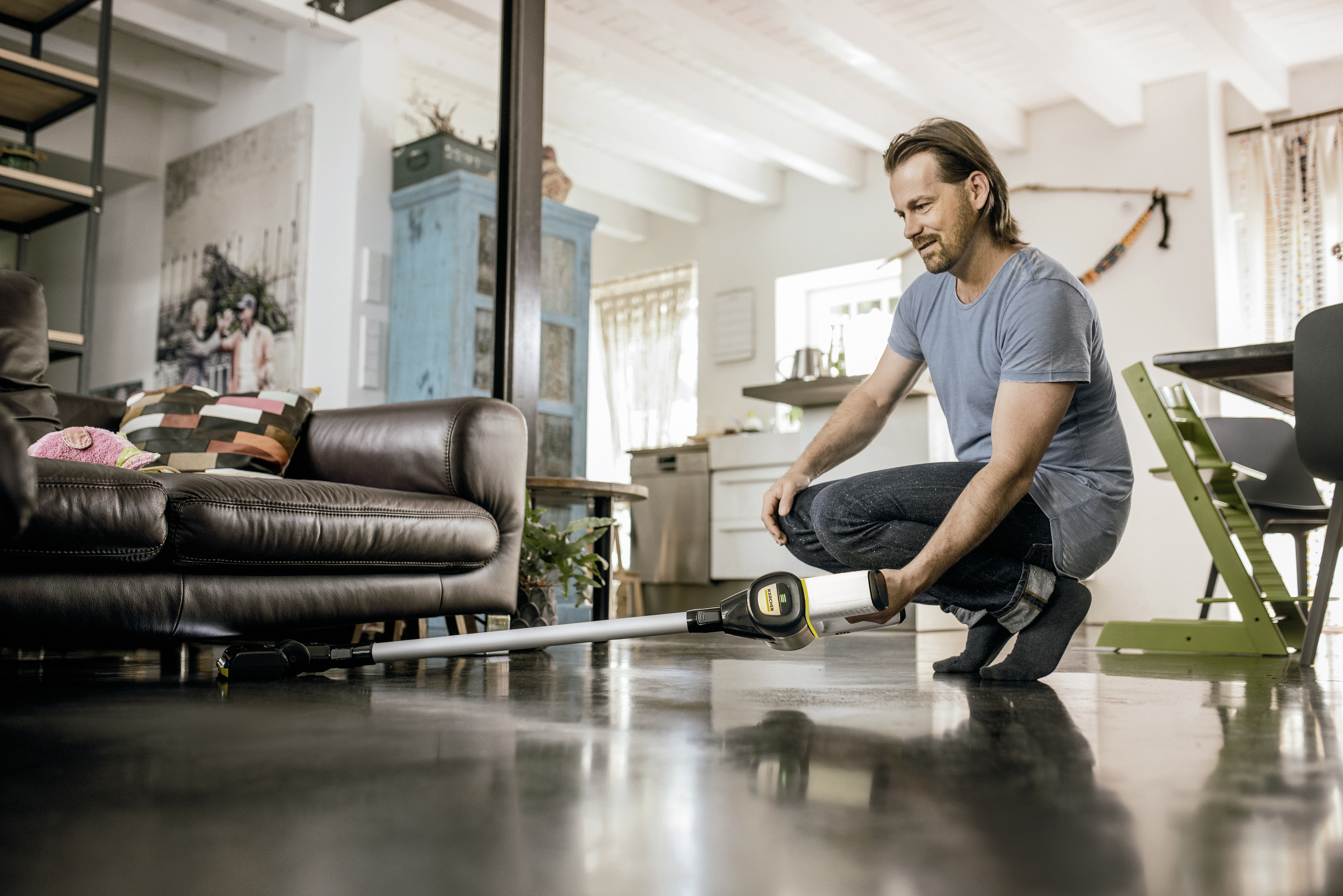 Man cleans floor with a cordless vacuum. Brown sofa and blue cabinet are in the background.