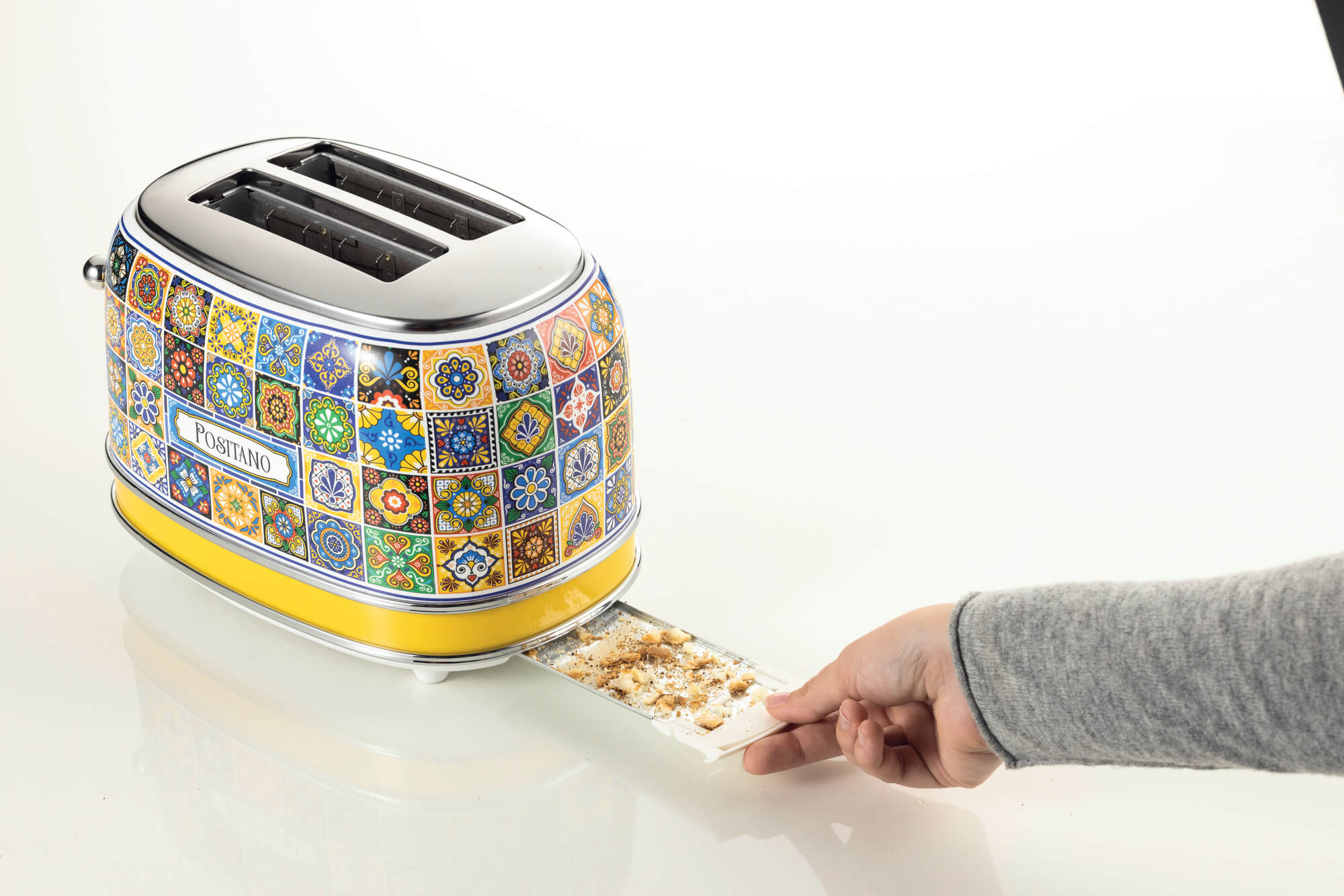 A colorful, patterned toaster with a hand removing bread crumbs from the tray on a white surface.