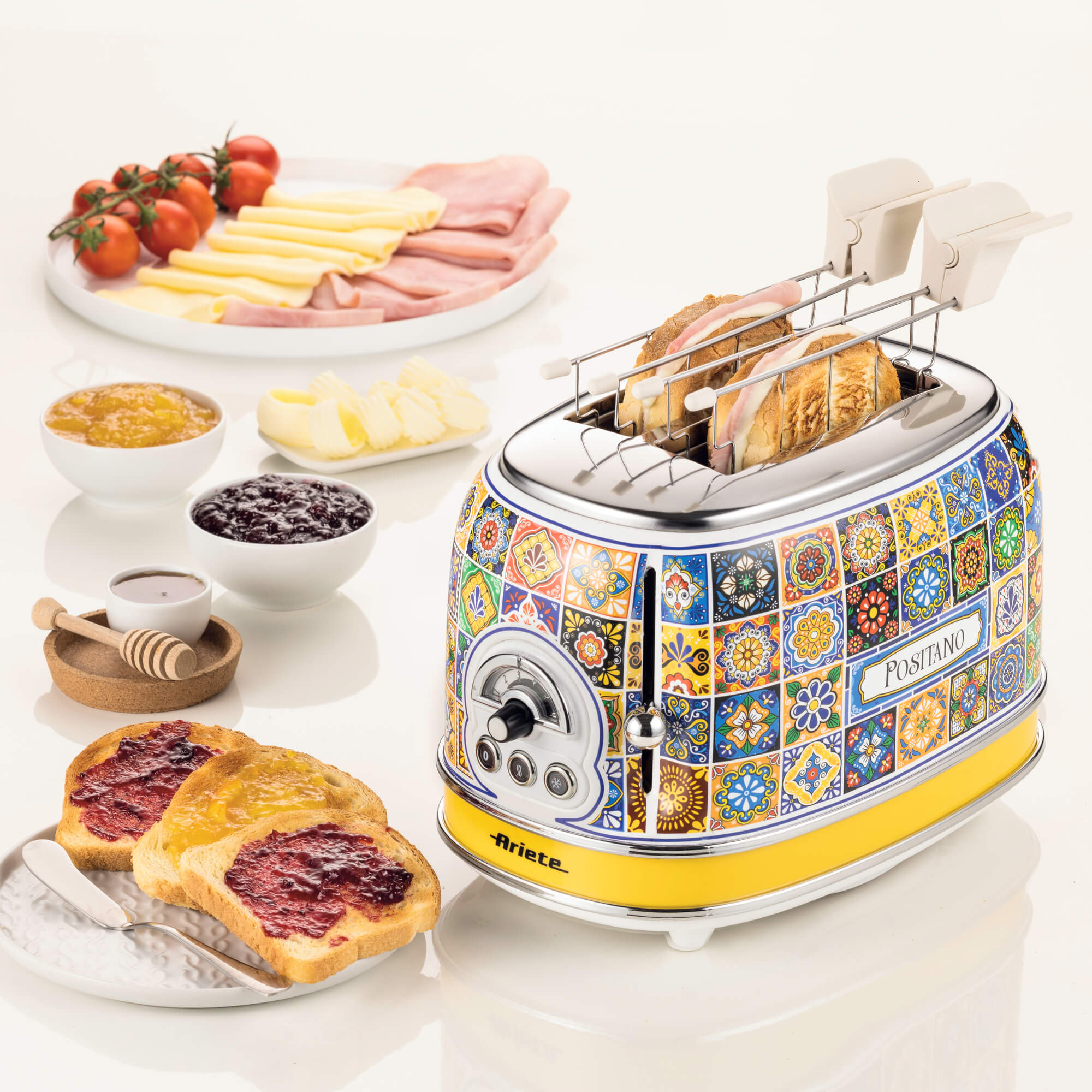 Toaster with bread, jam, butter, and tomatoes on a white surface.