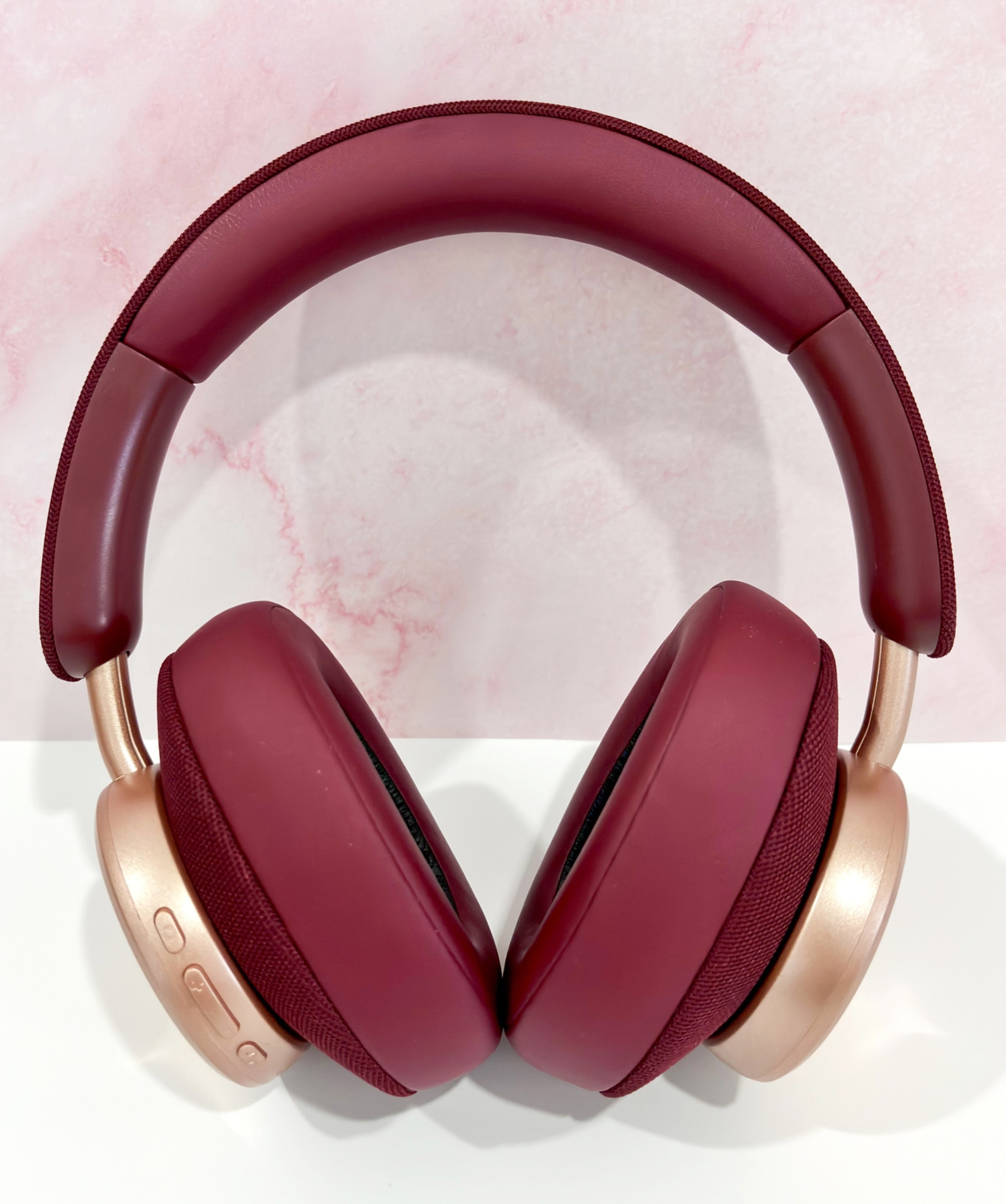 Auriculares borgoña sobre una superficie blanca, con fondo de mármol rosa. Los auriculares tienen detalles dorados.