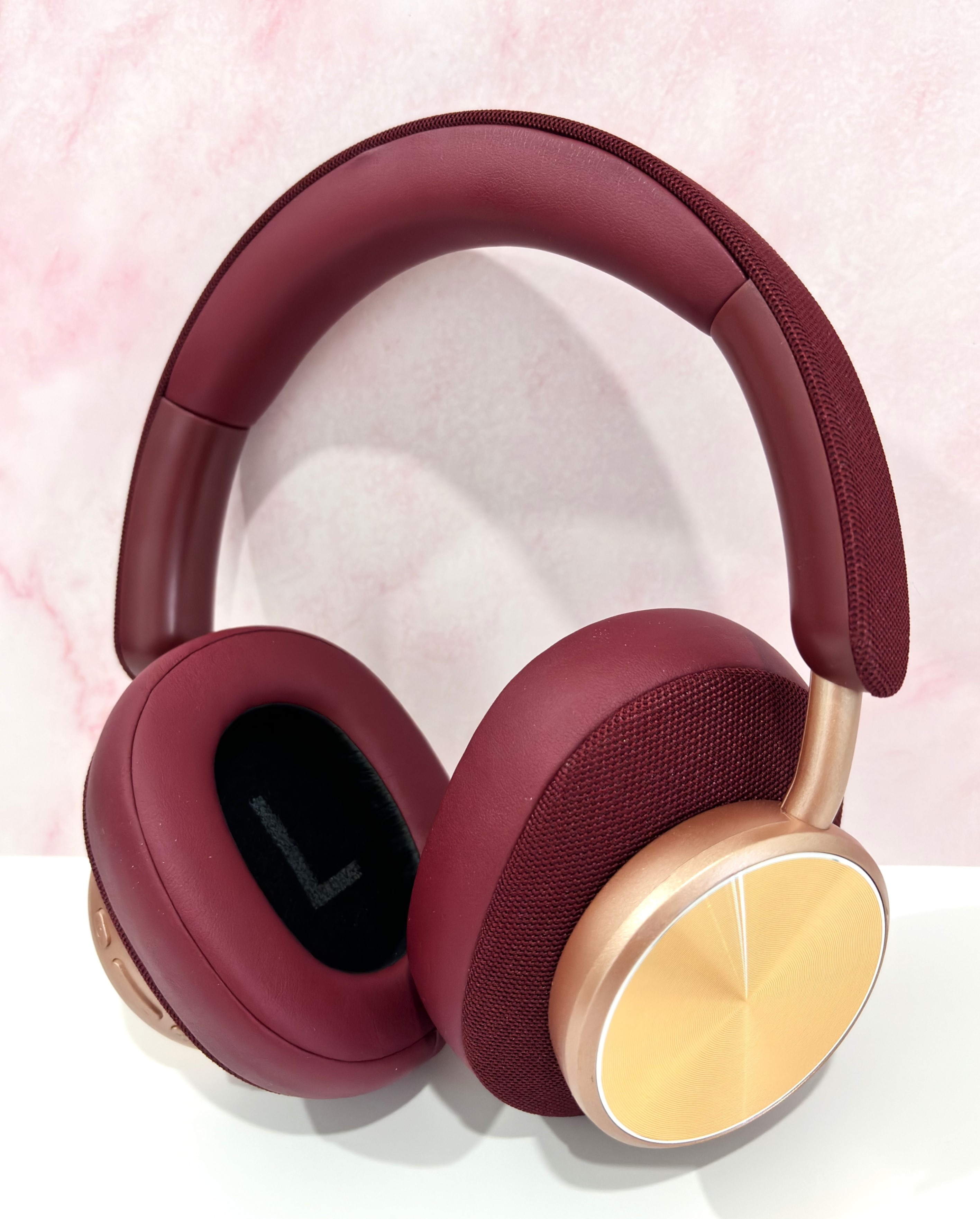 Auriculares color borgoña con detalles dorados. Los auriculares están sobre una superficie blanca con un fondo rosa.