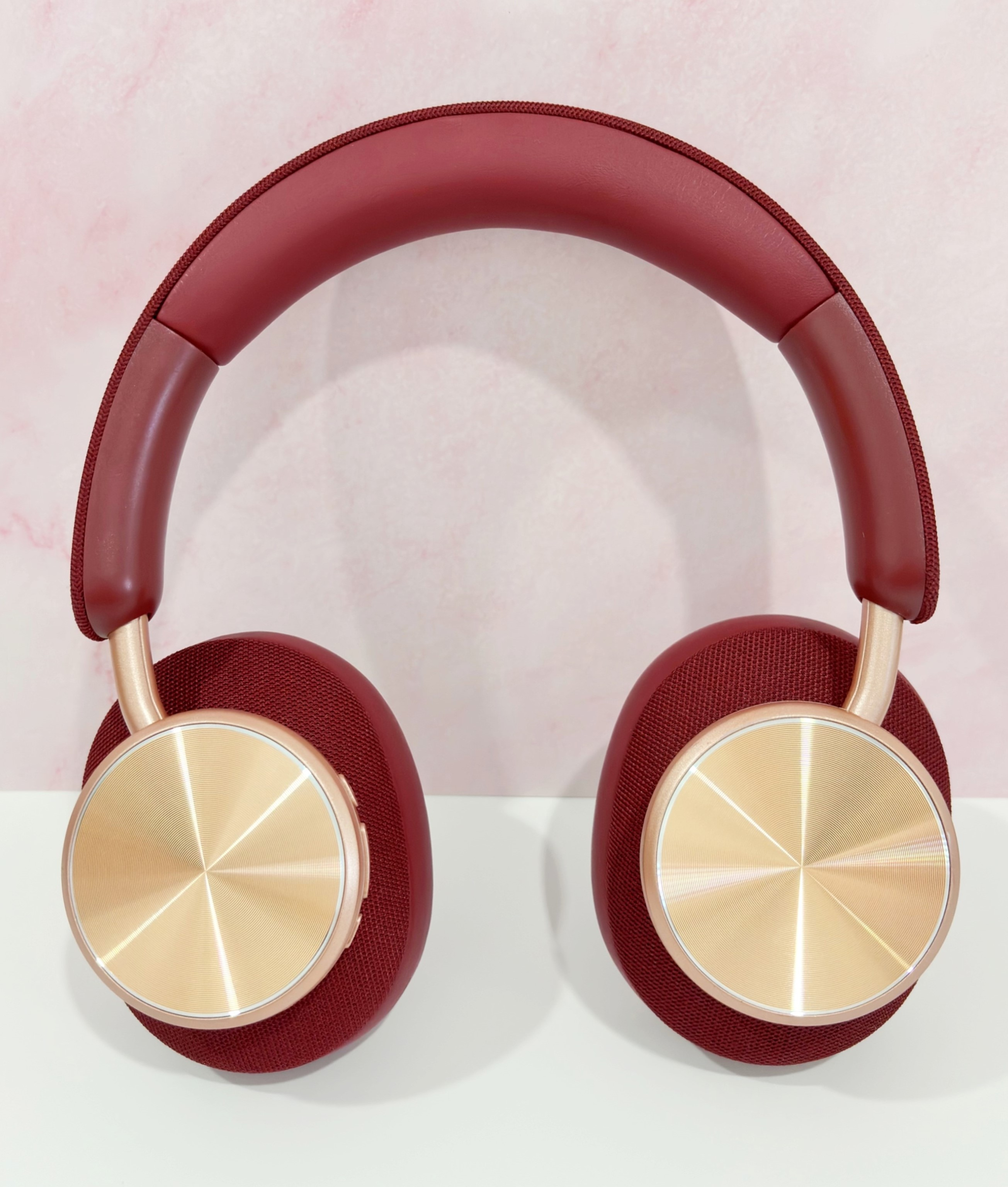 Auriculares burdeos con círculos dorados en las copas. La diadema también es burdeos. Fondo rosa.