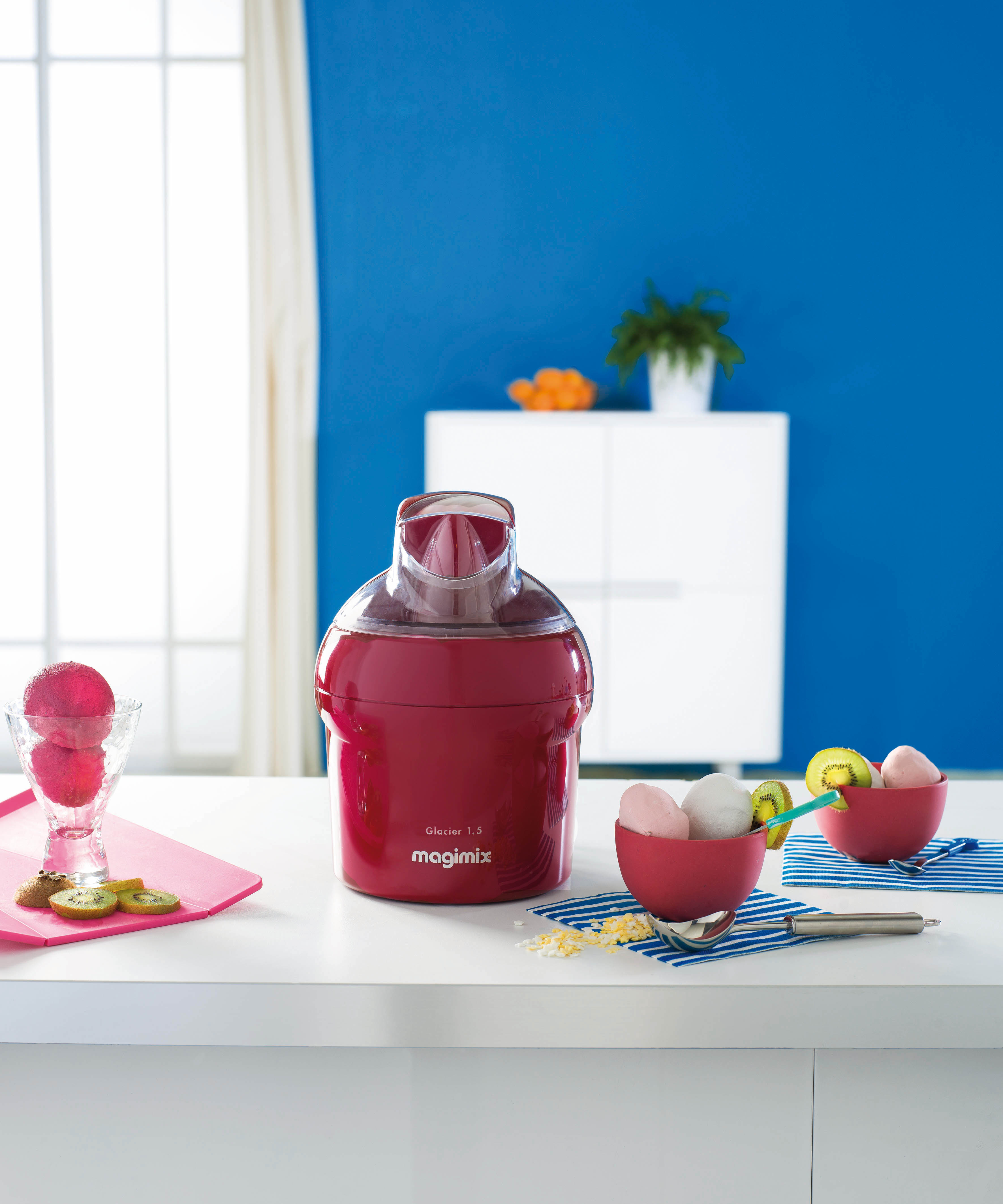 Machine à glace rouge sur une table blanche. Des bols de glace et de fruits sont à proximité, fond de mur bleu.