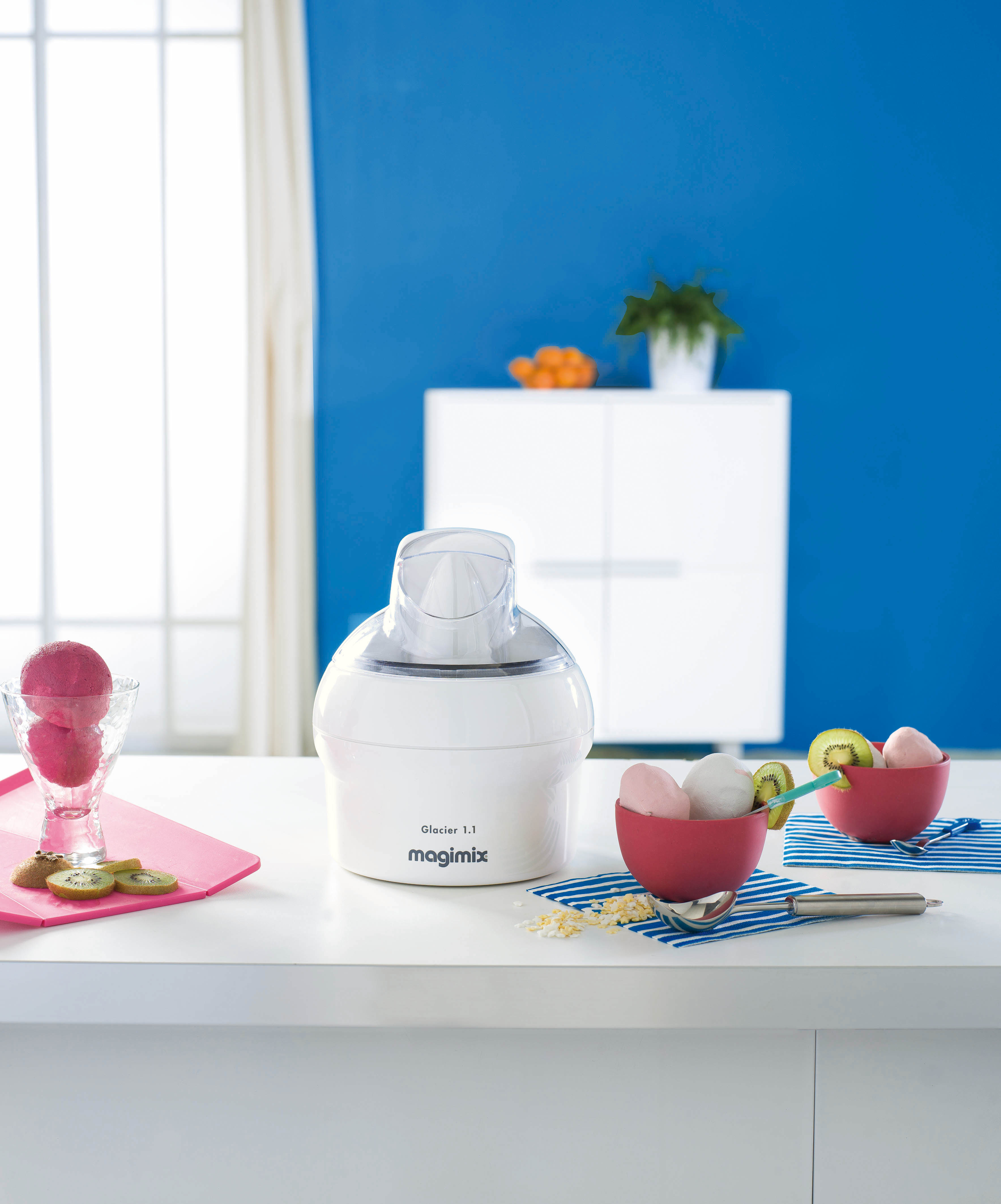 White ice cream maker with pink ice cream and kiwi slices on a white table, blue wall in background.