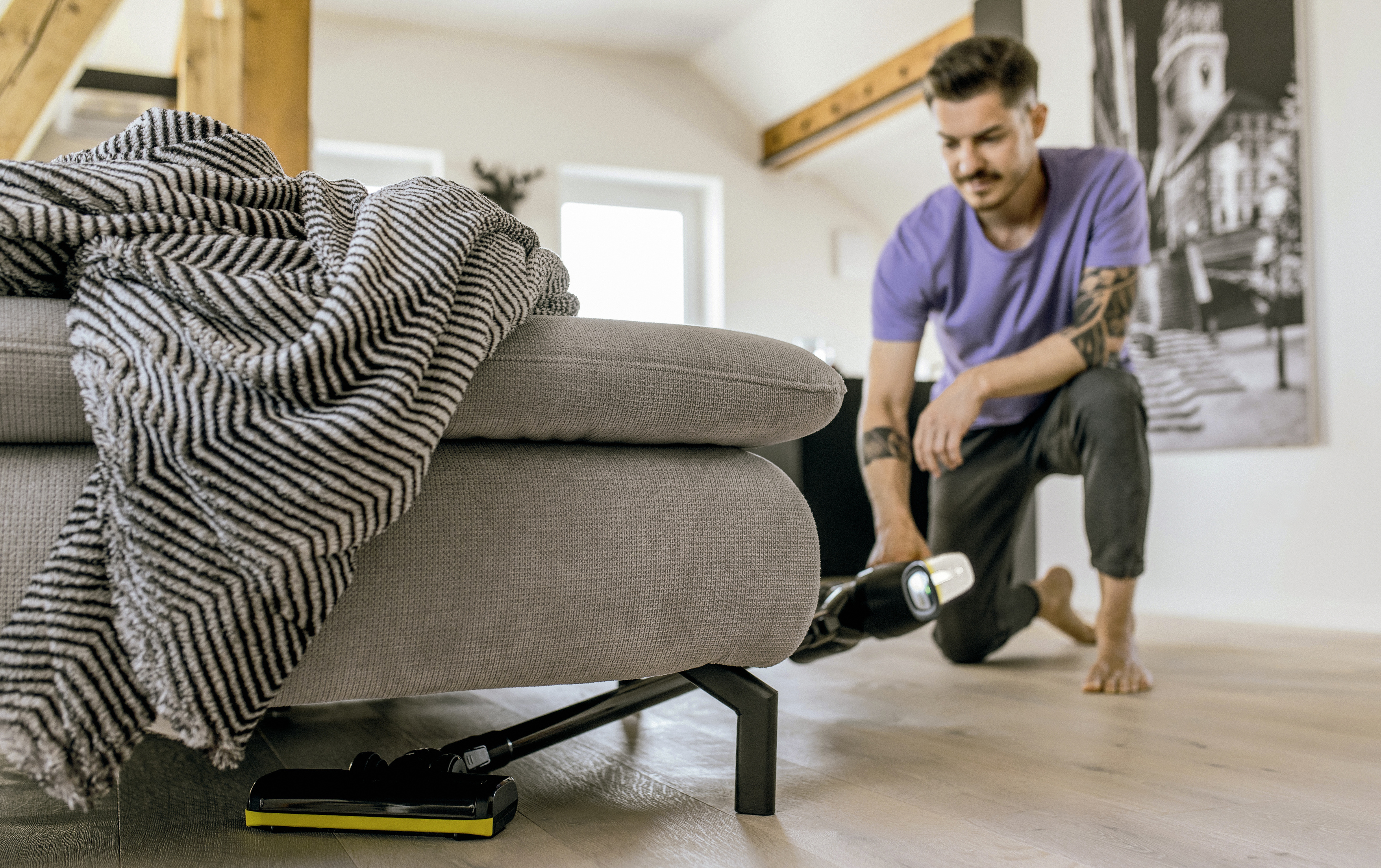 Une personne qui passe l'aspirateur sous un canapé gris. Une couverture est drapée sur le canapé.