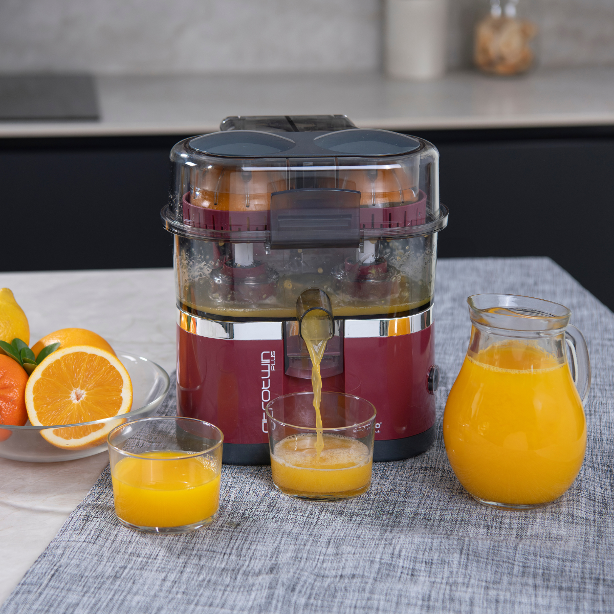 Una exprimidora con dos vasos de zumo y una jarra de zumo, junto a naranjas sobre una mesa.