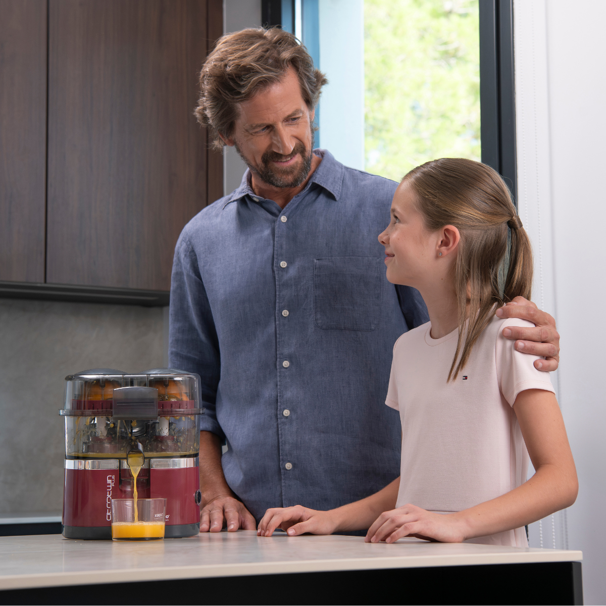 Hombre y niña usando una exprimidora. Electrodoméstico rojo y plateado. Ambiente de cocina.