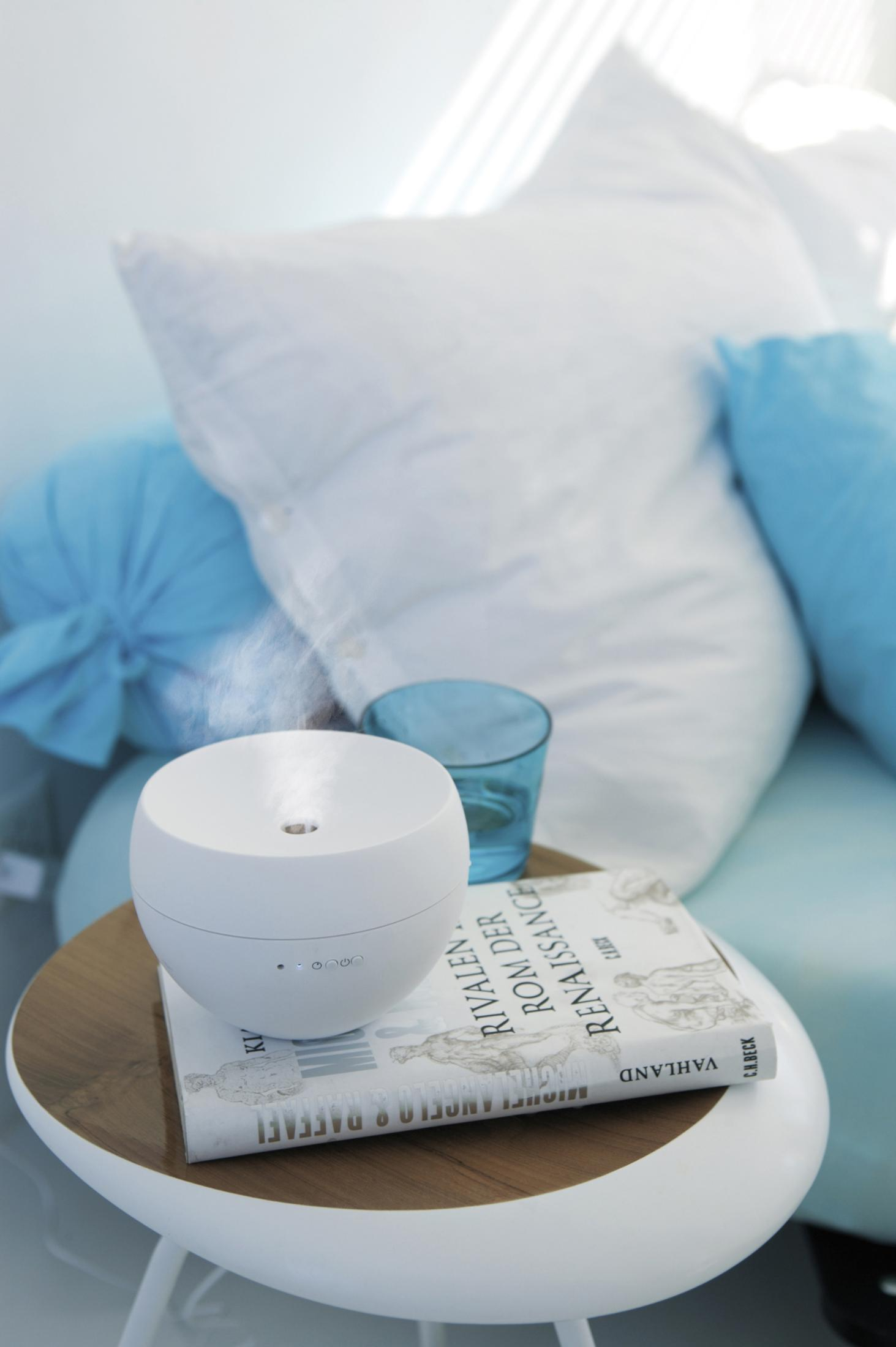 A white essential oil diffuser on a table with a book and a glass. Pillows in the background.