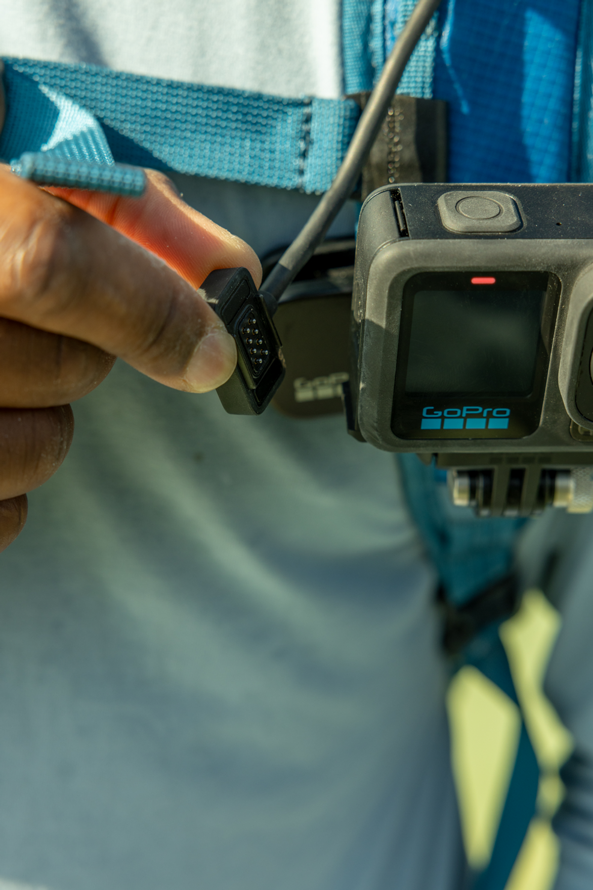 Mano che collega un cavo nero a una fotocamera GoPro su un'imbracatura blu.