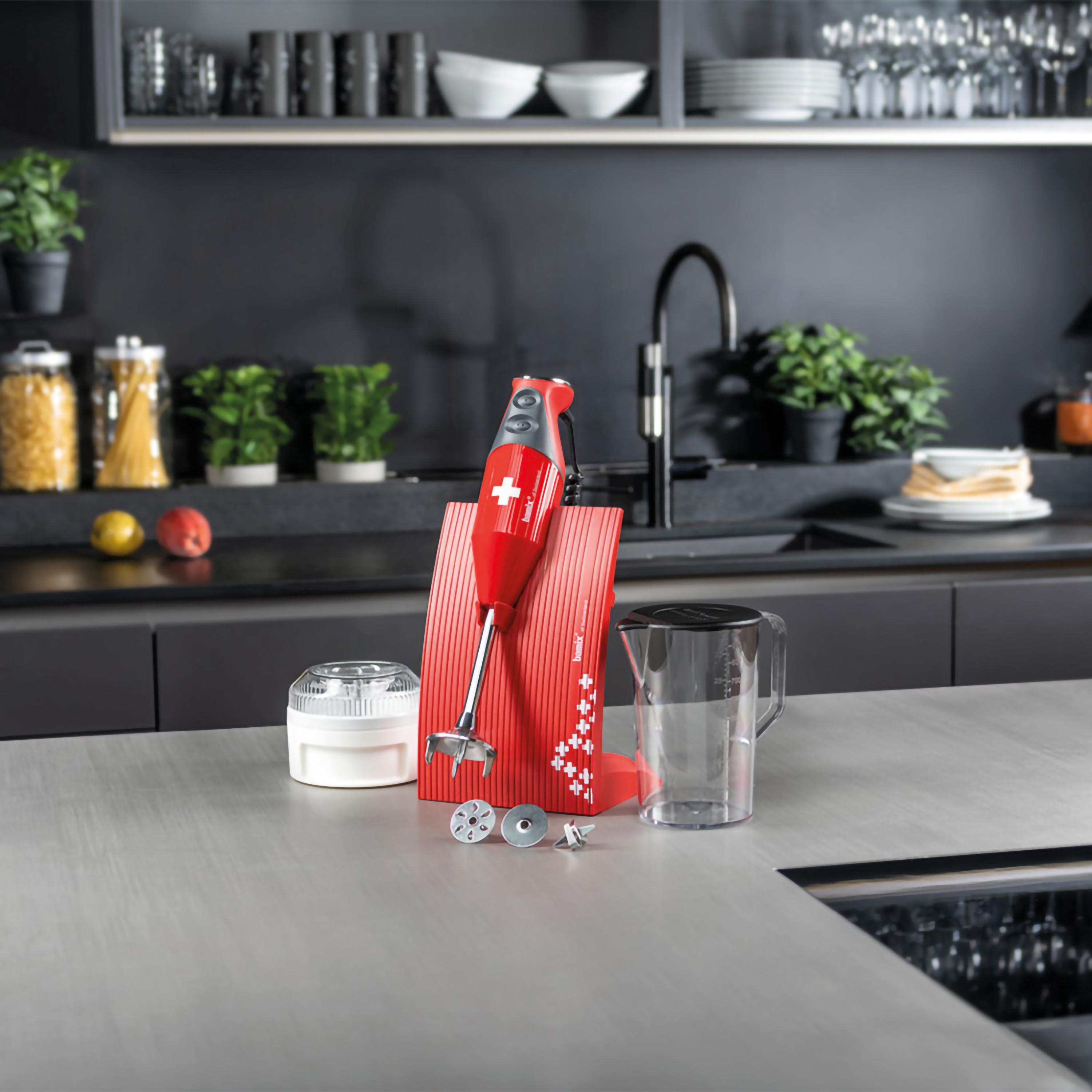 Un frullatore a immersione rosso con accessori, tra cui un supporto, posto su un piano cucina chiaro.