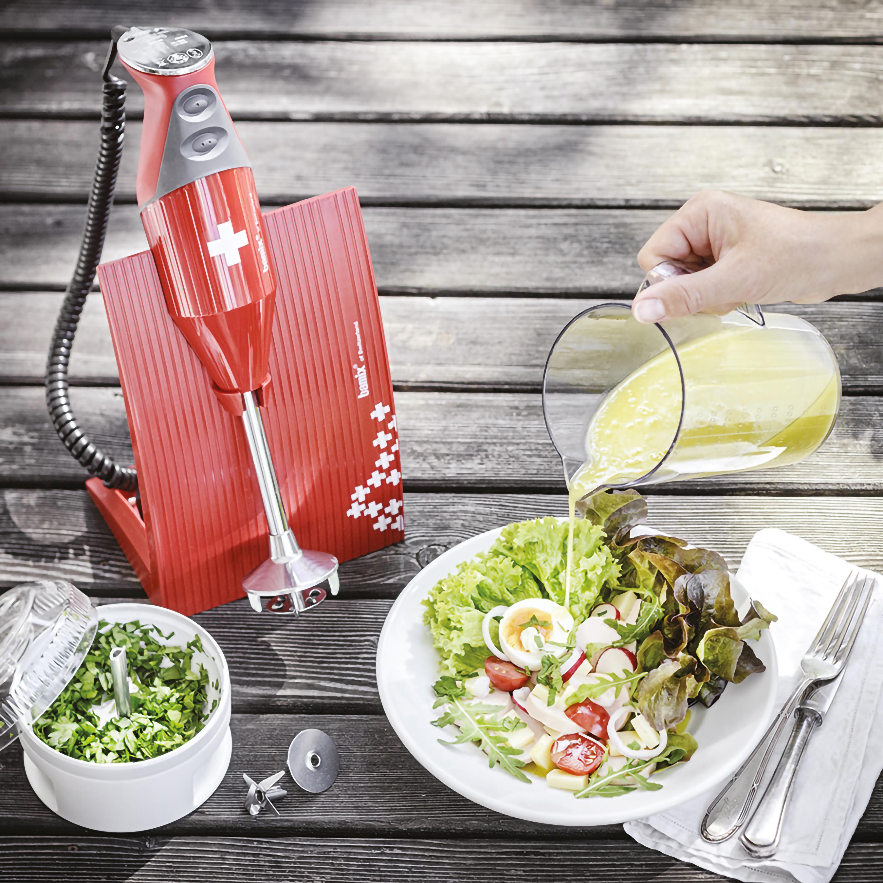 Persona versa condimento su insalata, frullatore rosso e accessori su tavolo in legno.