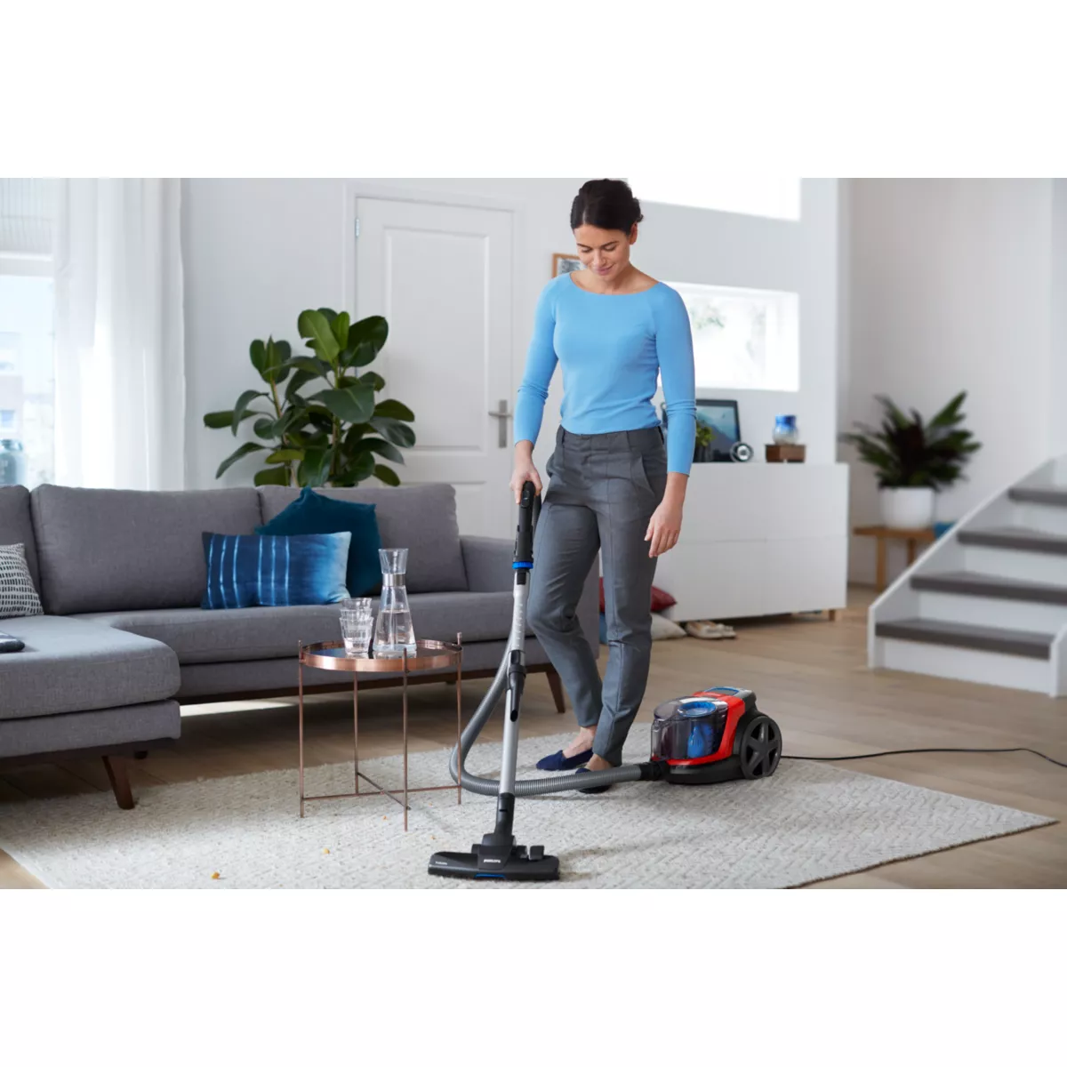 Kobieta odkurza dywan czerwono-niebieskim odkurzaczem w salonie.