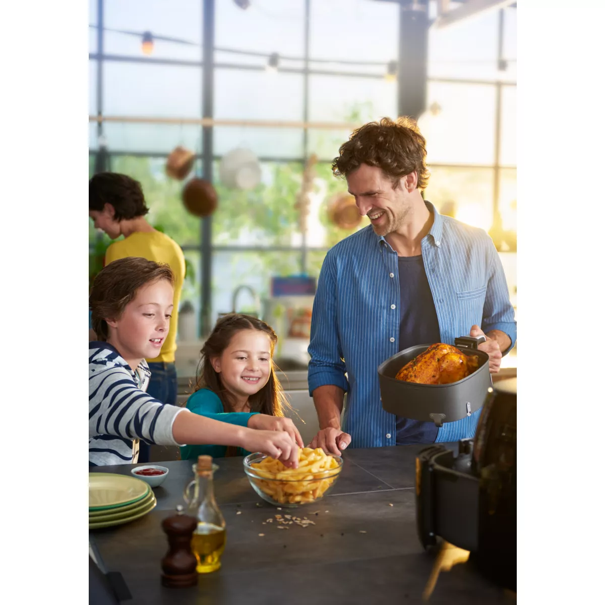 Una familia se reúne alrededor de una freidora de aire. El padre sostiene la freidora. Los niños están cerca de papas fritas y un cuenco.