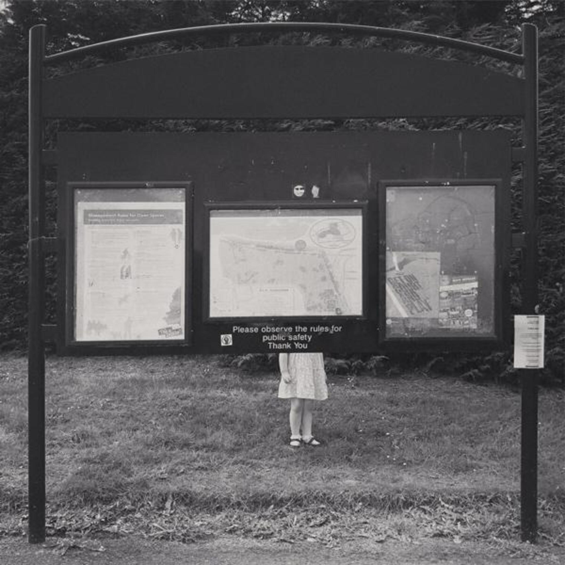 Une image en noir et blanc. Un panneau avec trois cadres d'informations, une fille se tient devant, dans l'herbe.