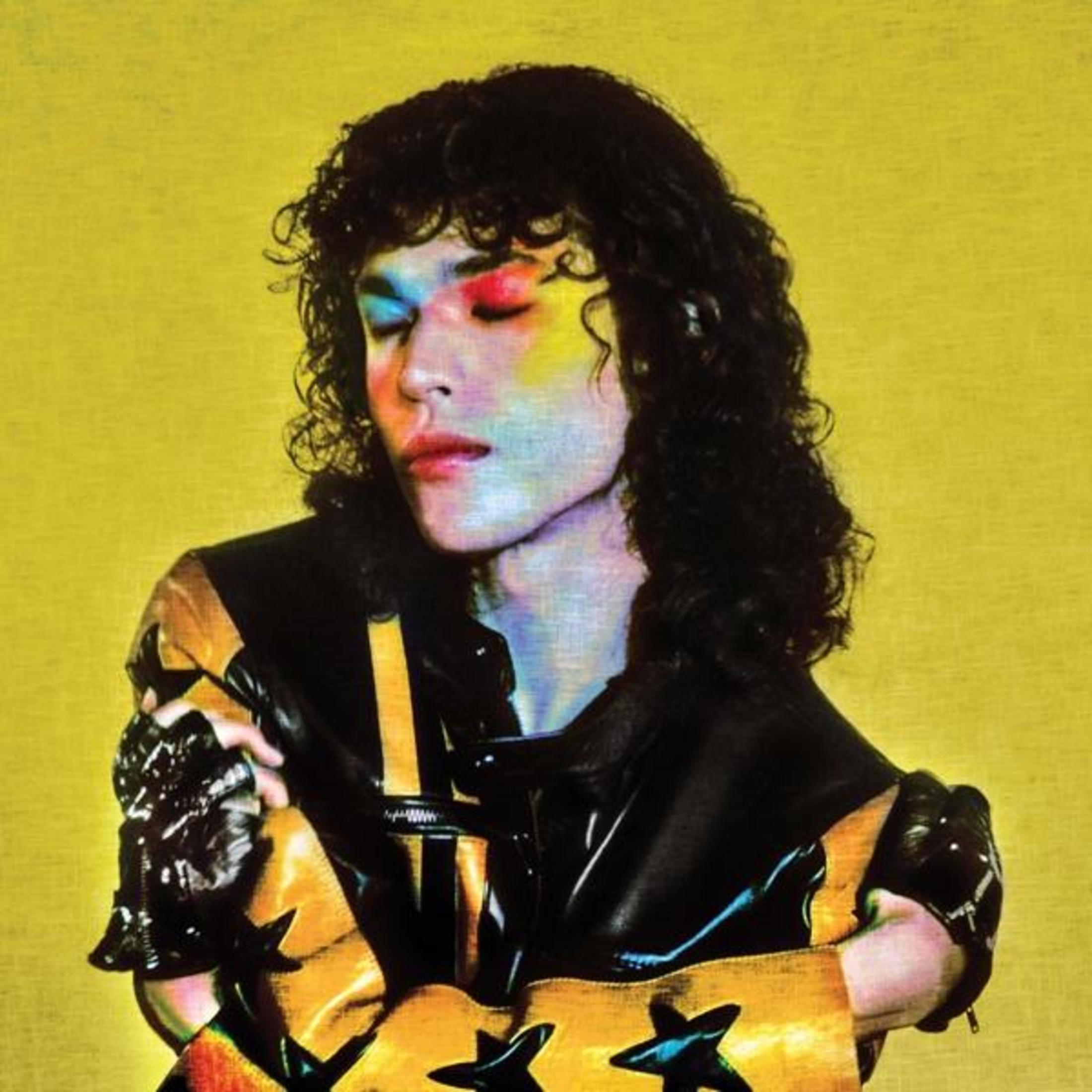 Persona con trucco colorato e capelli neri. Sfondo giallo. Indossa una giacca nera e oro.