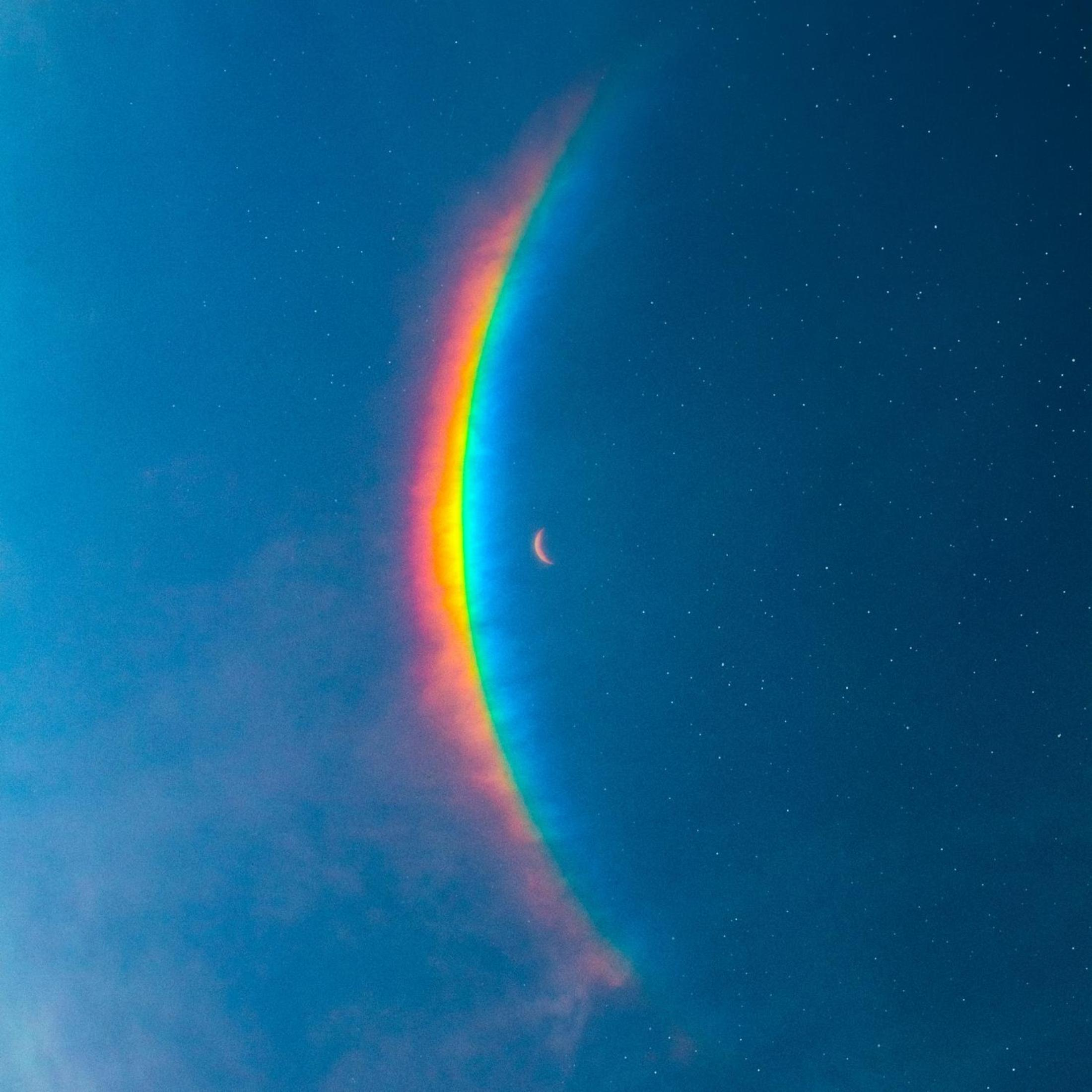 Een regenboog over een sterrenhemel, met een maansikkel rechts van de regenboog.