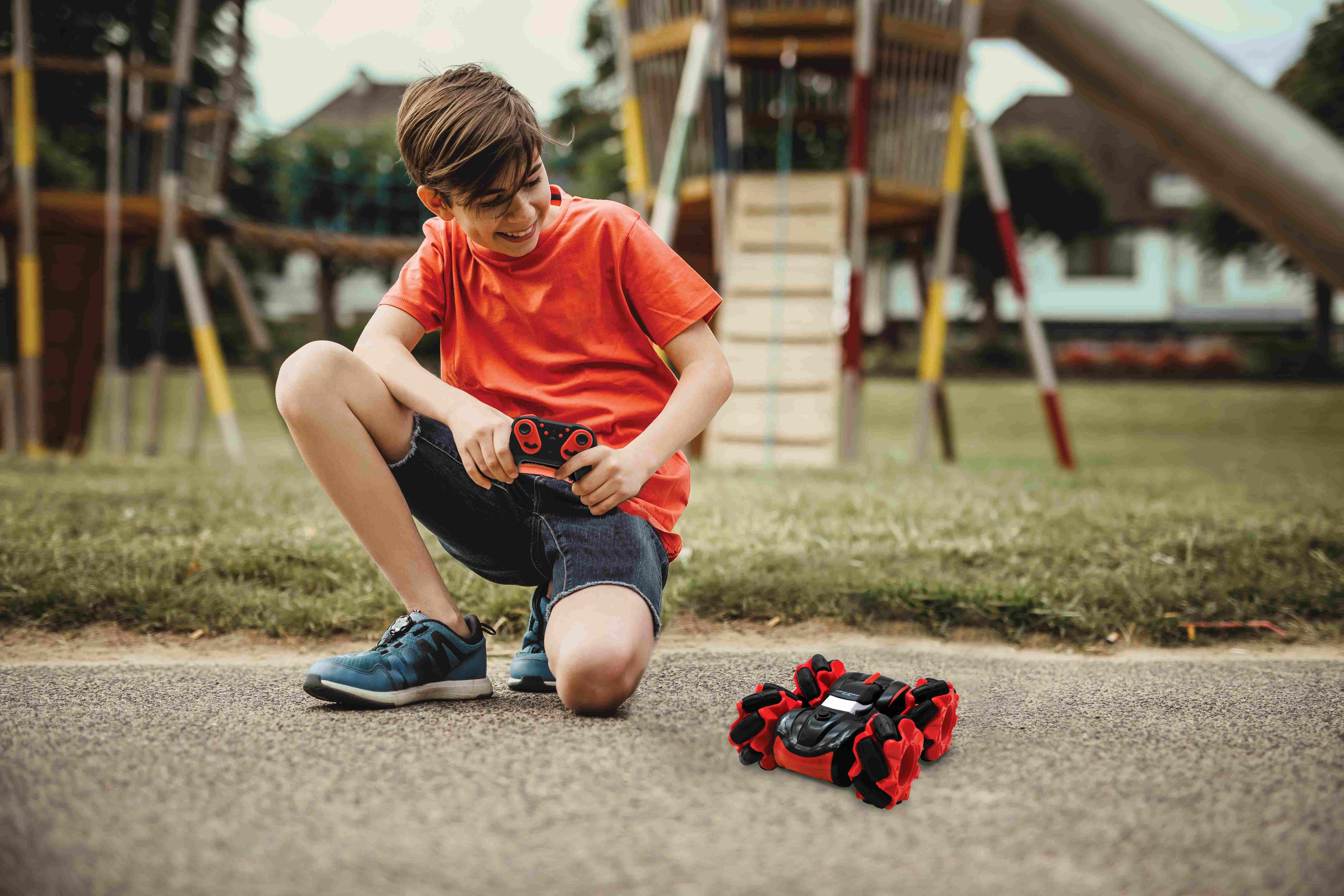 Niño con camiseta roja juega con un coche de juguete rojo y negro. Parque infantil al fondo.