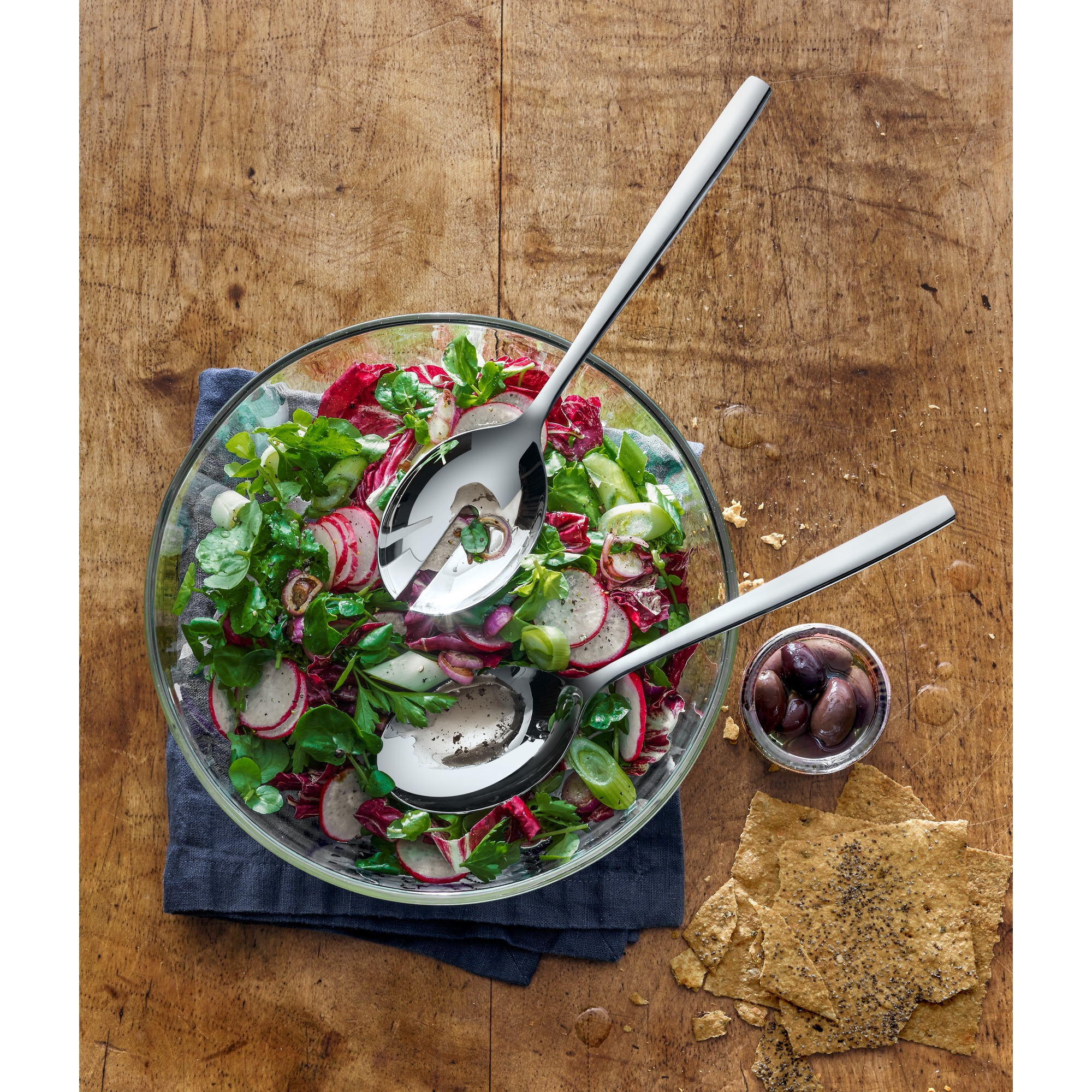 Salat in einer Glasschüssel mit Servierlöffeln und Oliven.
