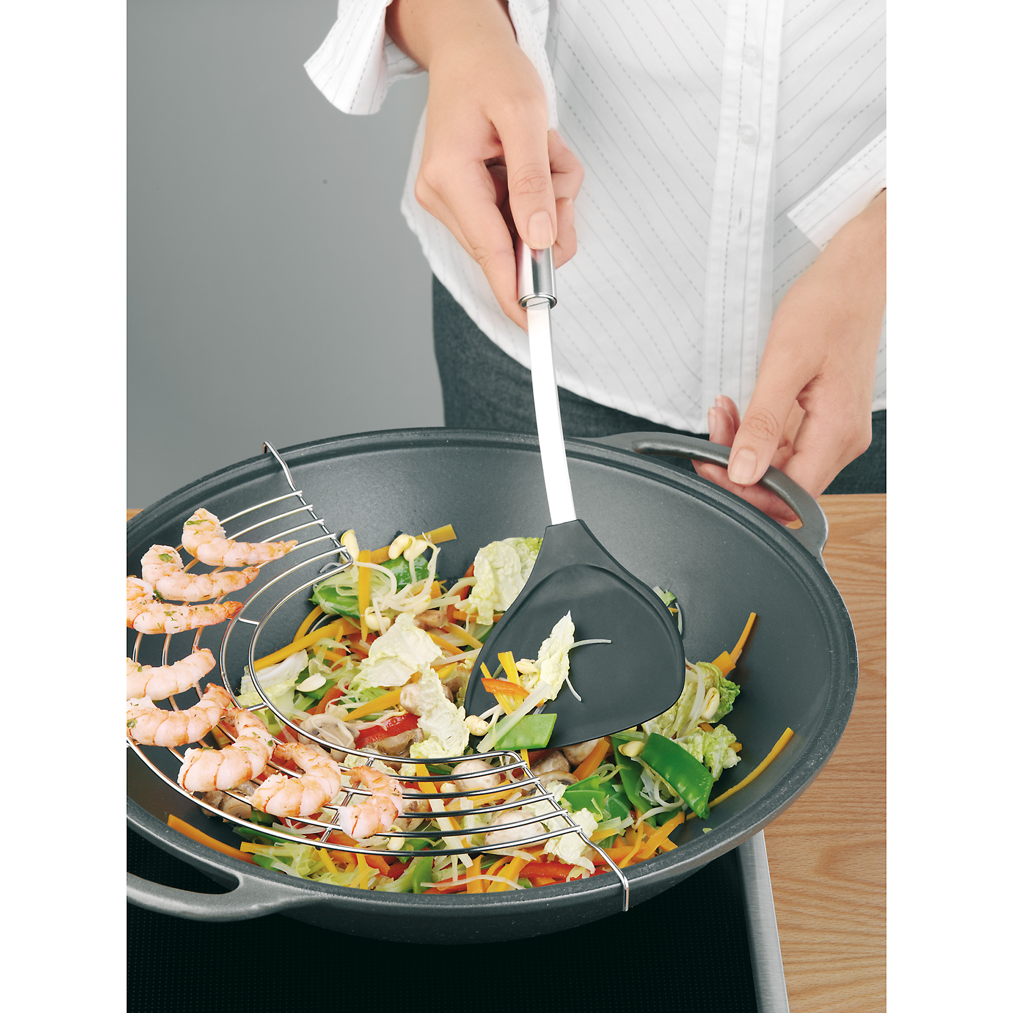 Osoba używa szpatułki do mieszania warzyw i krewetek w woku z parowarem.