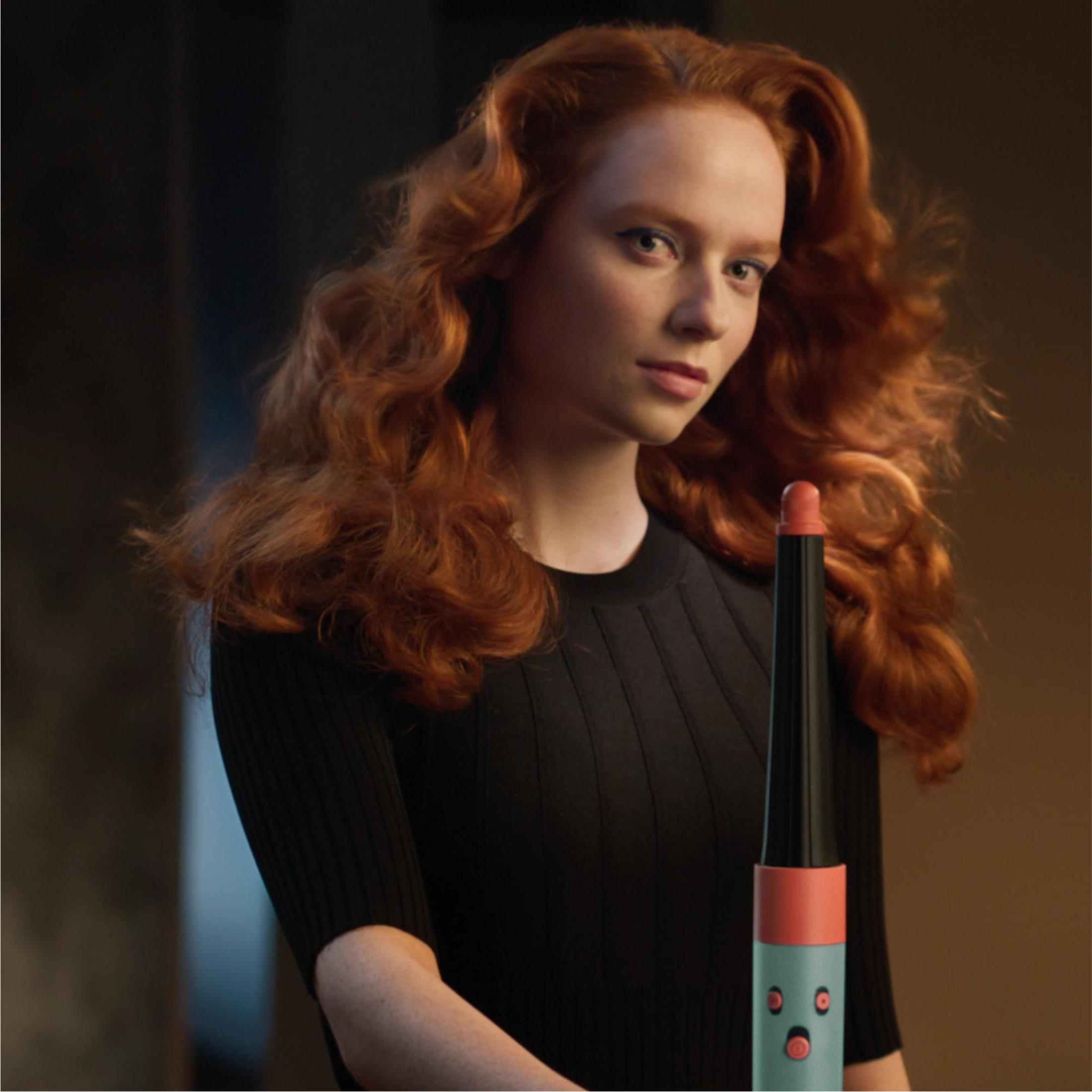 Kobieta z rudymi włosami trzyma lokówkę. Tło jest rozmyte i ma ciepły kolor.