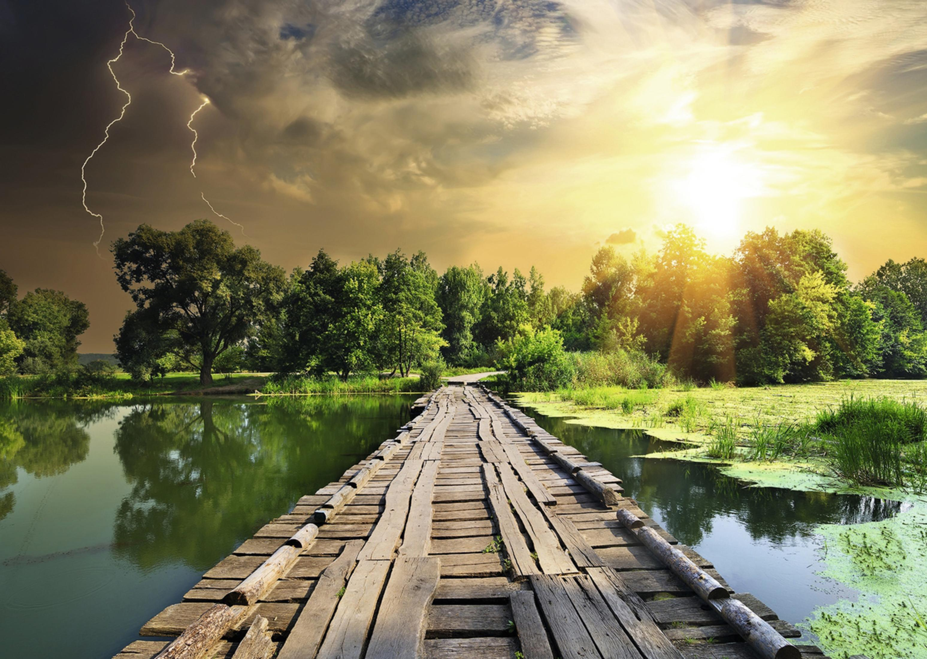 Eine lange Holzbrücke über Wasser, Bäume an den Ufern, ein sonniger Himmel mit dunklen Wolken und Blitz.