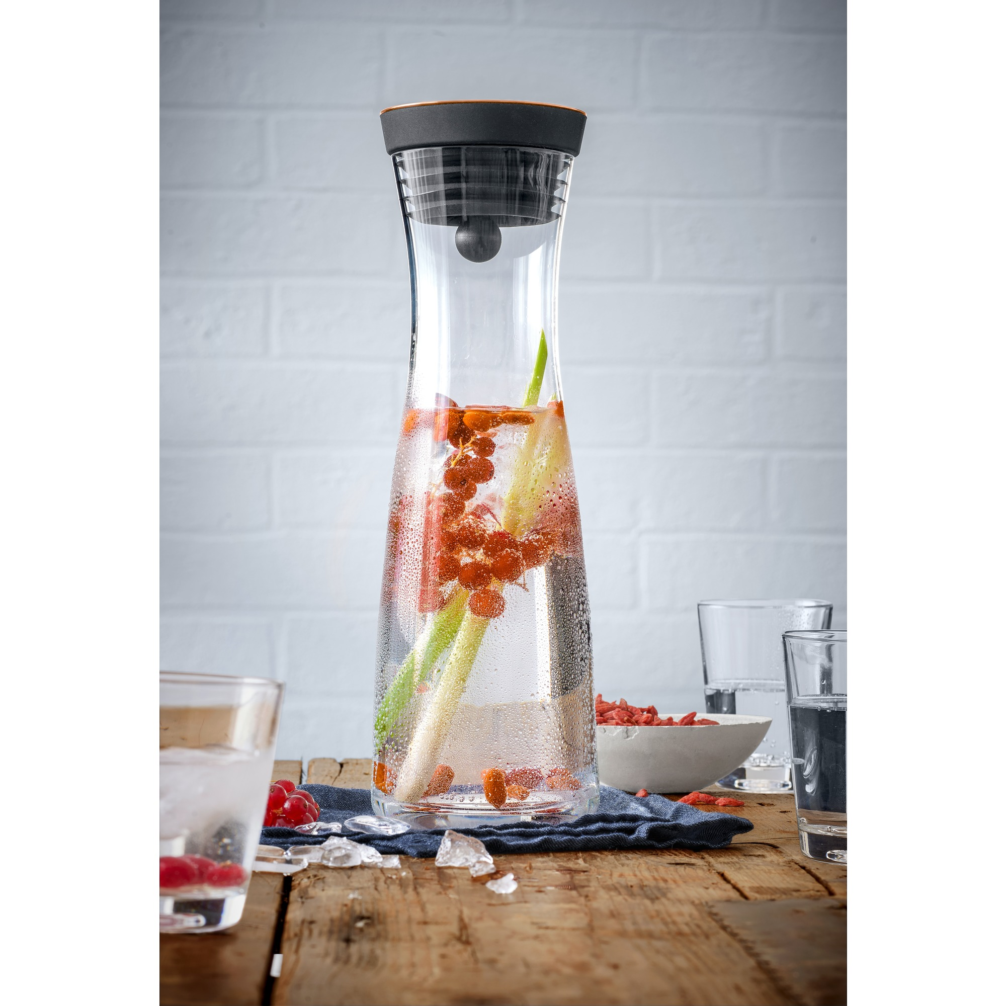 Ein klarer Glaskrug mit Wasser, Früchten und Eis. Holztisch und Gläser im Hintergrund.