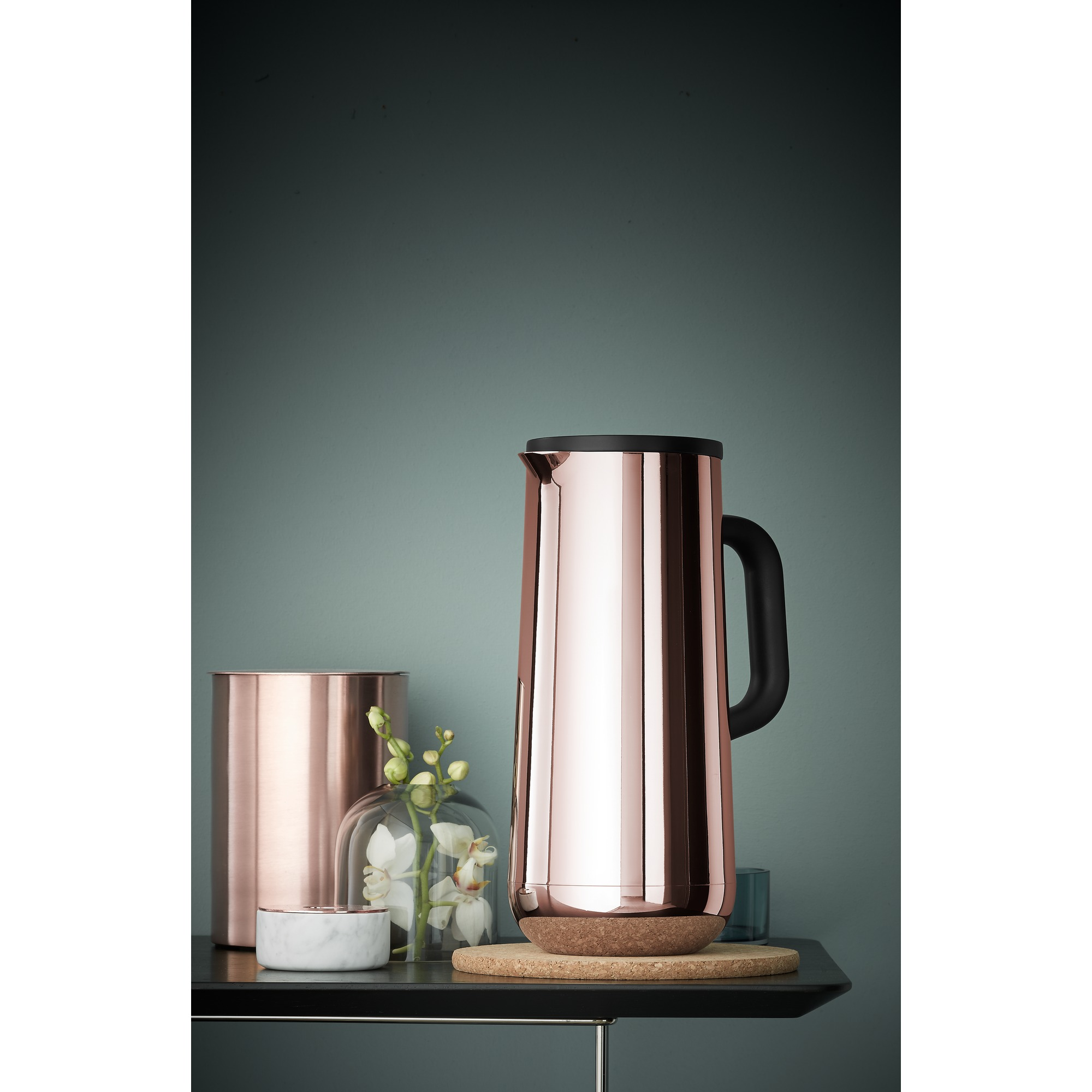Un pichet cuivré et un vase avec des fleurs sont posés sur une surface sombre. Un dôme en verre est également visible.