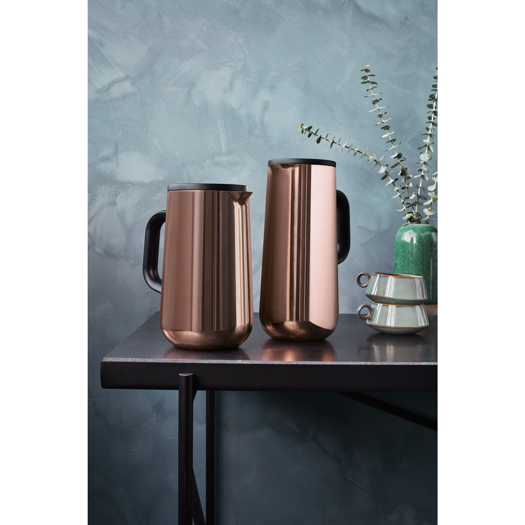 Deux carafes thermiques cuivrées. Une table et un vase avec des plantes. Fond bleu.