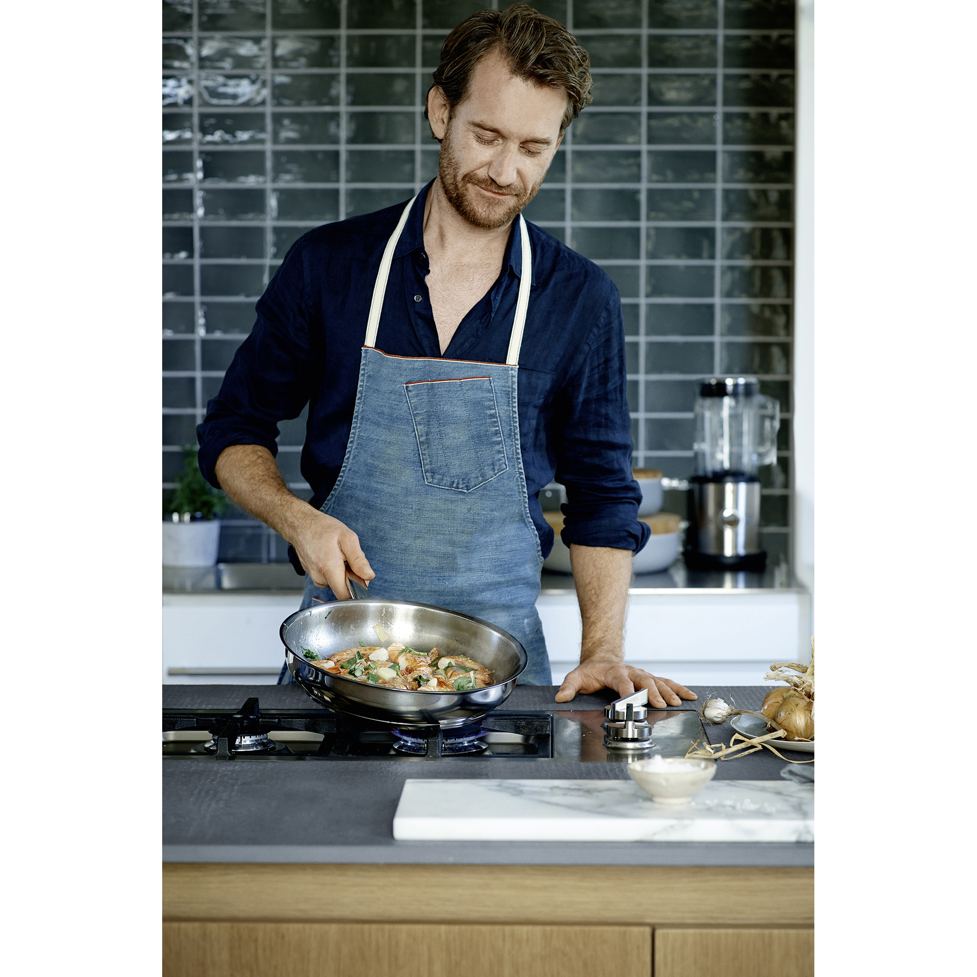 Homme cuisinant dans une poêle sur une cuisinière. Portant un tablier bleu. Légumes visibles.