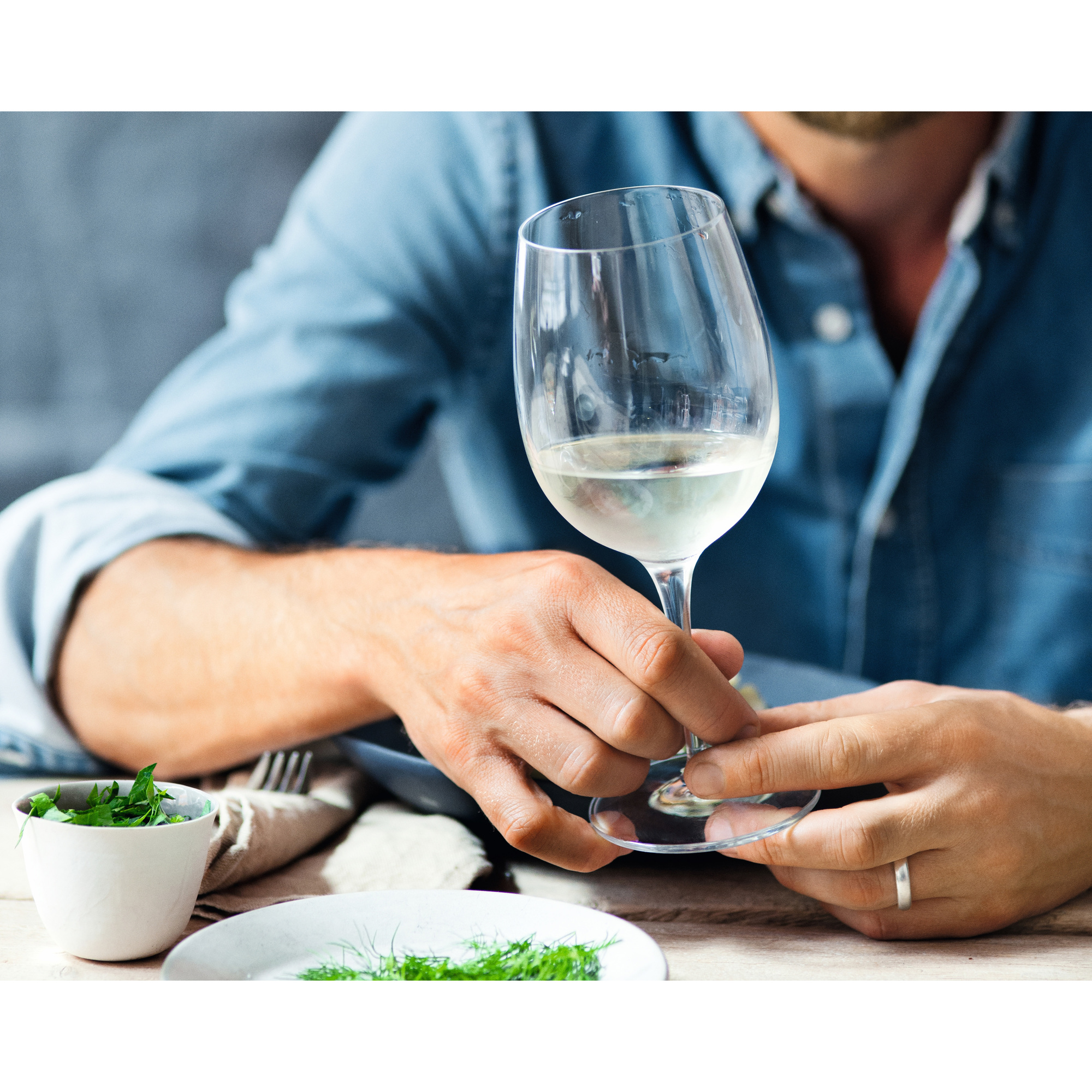 Eine Person, die ein Glas Weißwein hält. Eine kleine Schüssel mit grünen Kräutern und ein weißes Tuch liegen auf dem Tisch.