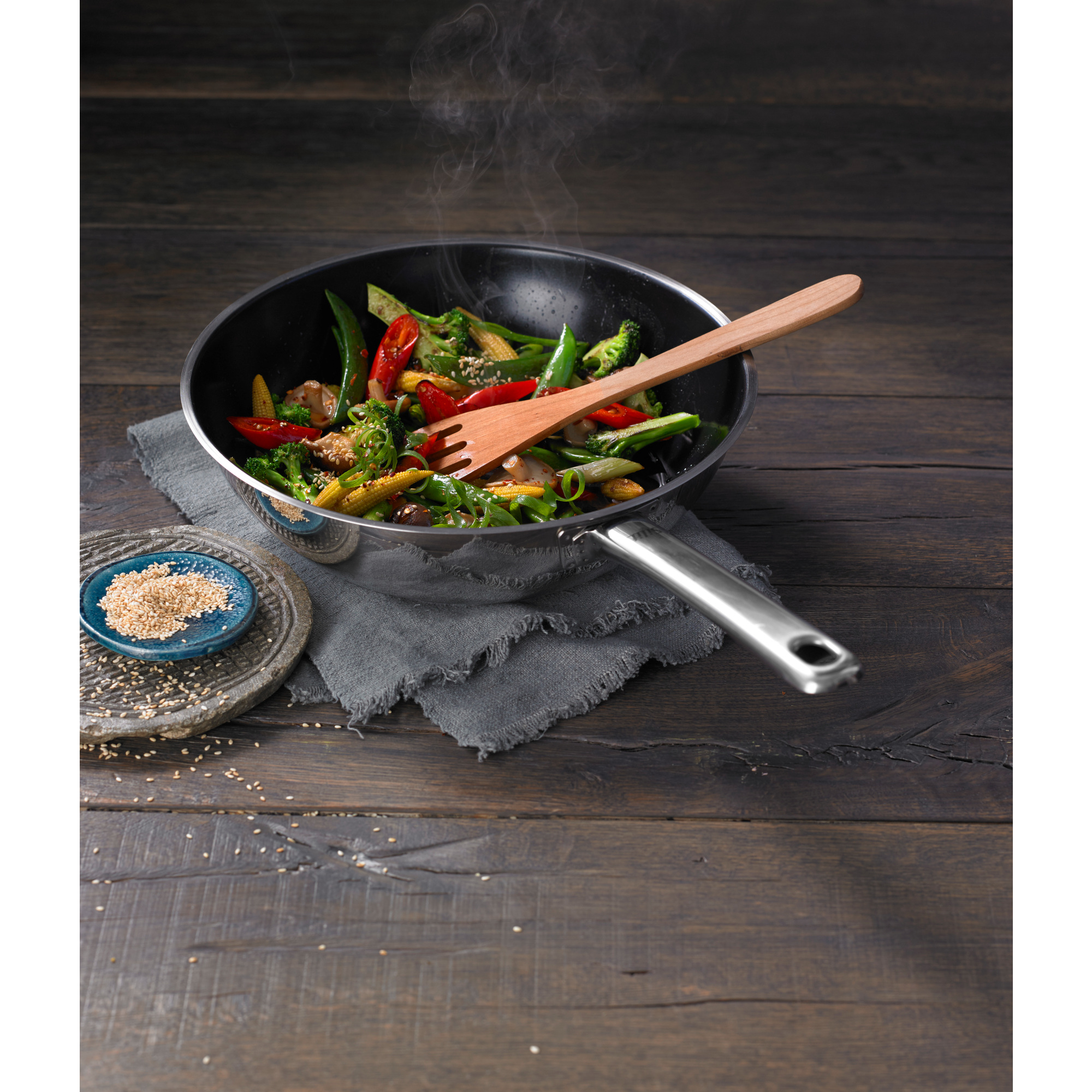 Ein Wok mit Gemüse und einer hölzernen Gabel, mit einem Sesamteller auf einem dunklen Holztisch.