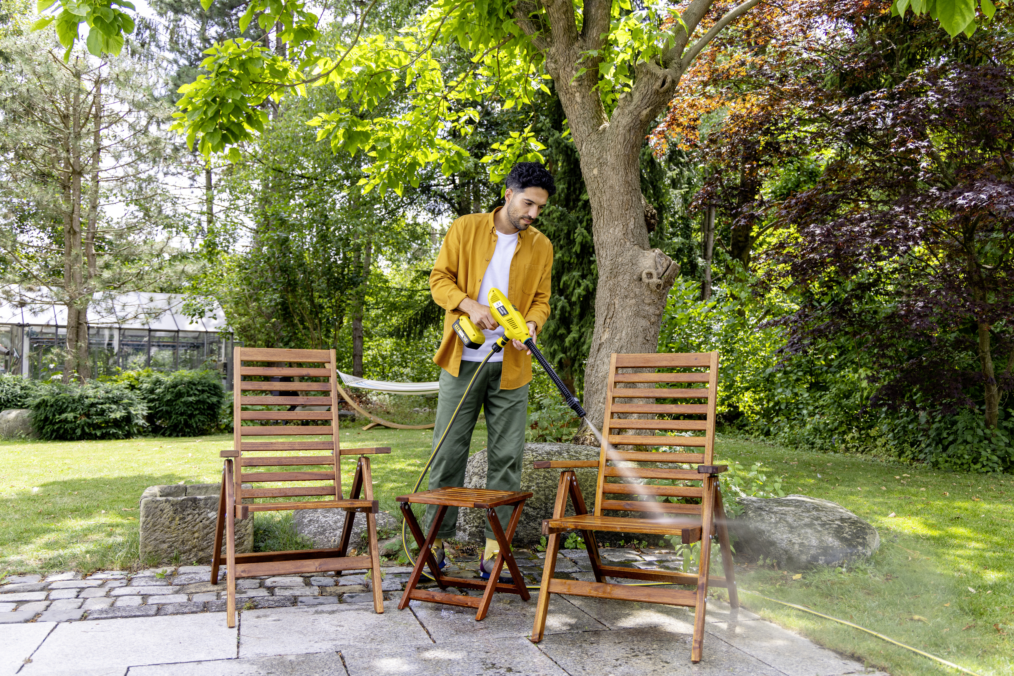 A man cleaning chairs with a power washer in a sunny garden with trees and a hammock.