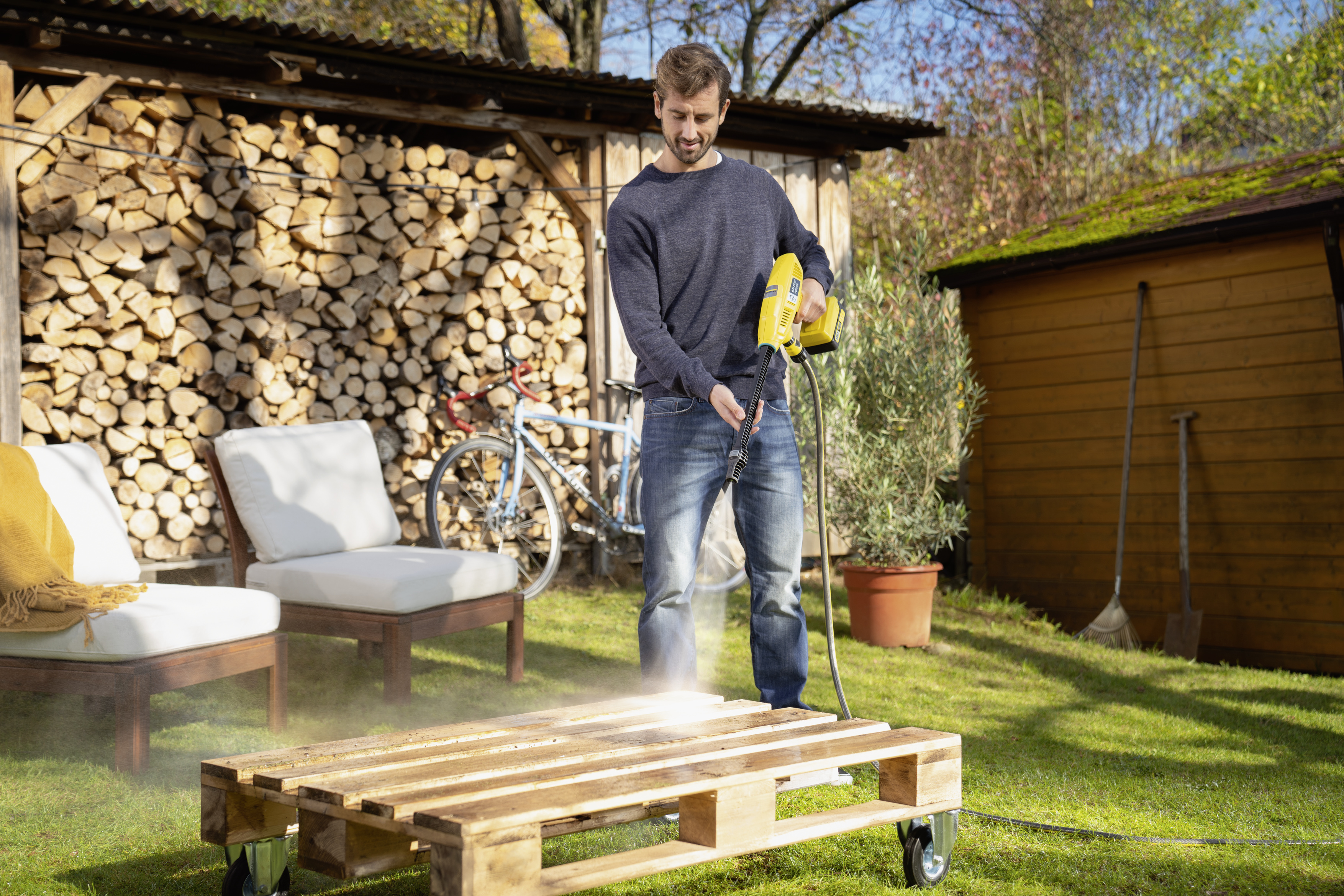 Man using a pressure washer on a wooden pallet. Backyard scene with firewood and a shed.