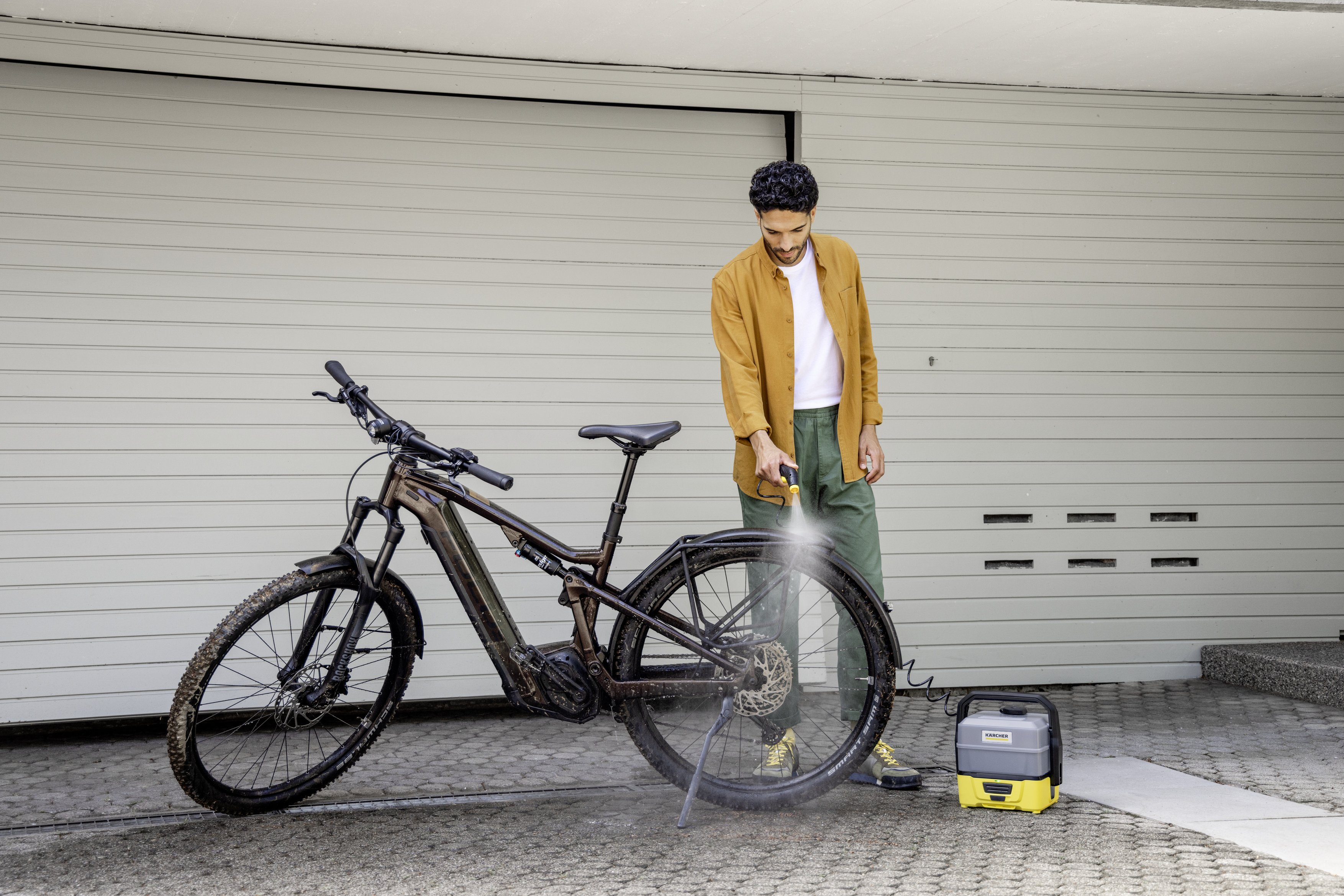 Un homme lave un vélo avec un nettoyeur haute pression, le vélo est devant une porte de garage.