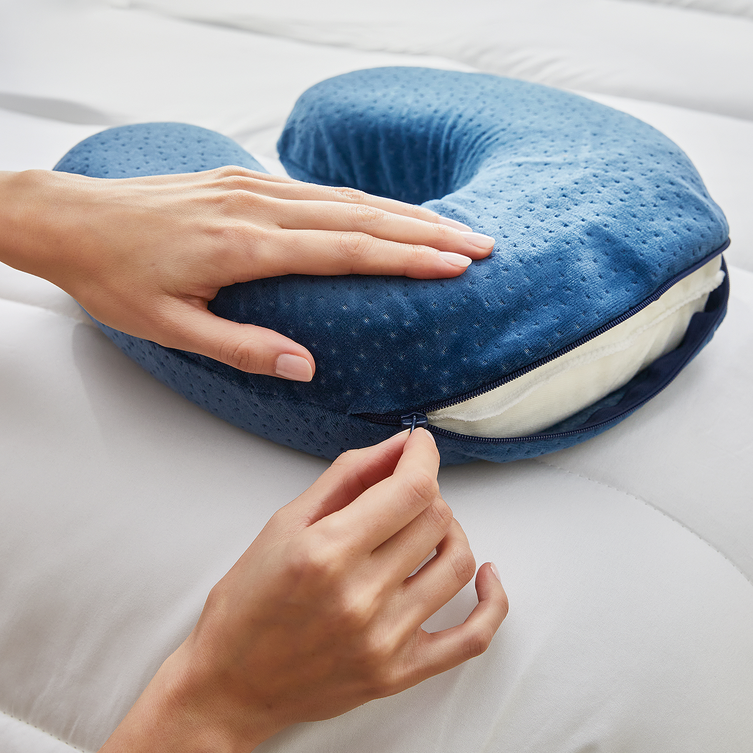 Manos cerrando una almohada de viaje azul en una cama blanca. Fondo crema.
