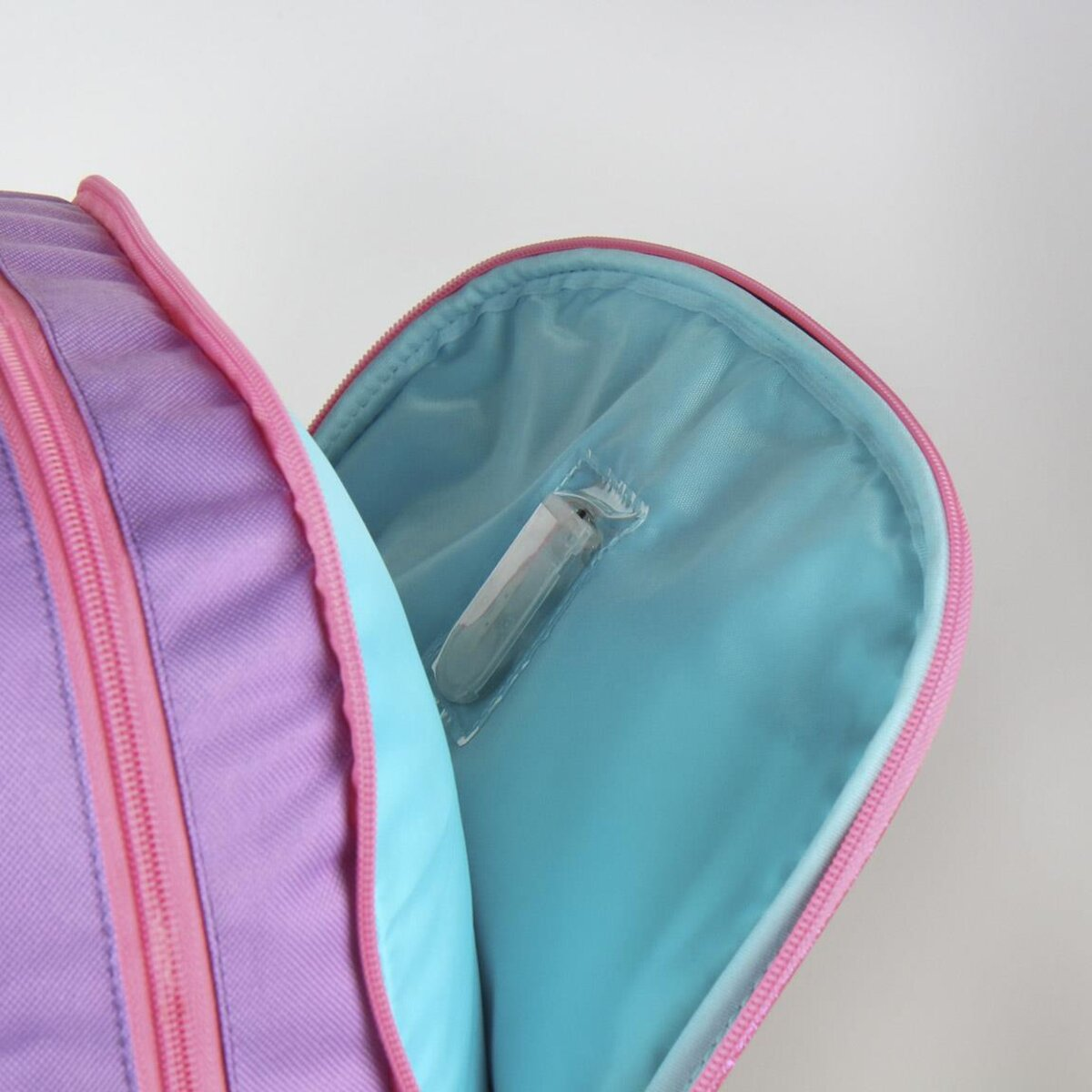 Una mochila morada con cremallera rosa está abierta, revelando un interior azul claro. Un objeto envuelto en transparente está dentro.