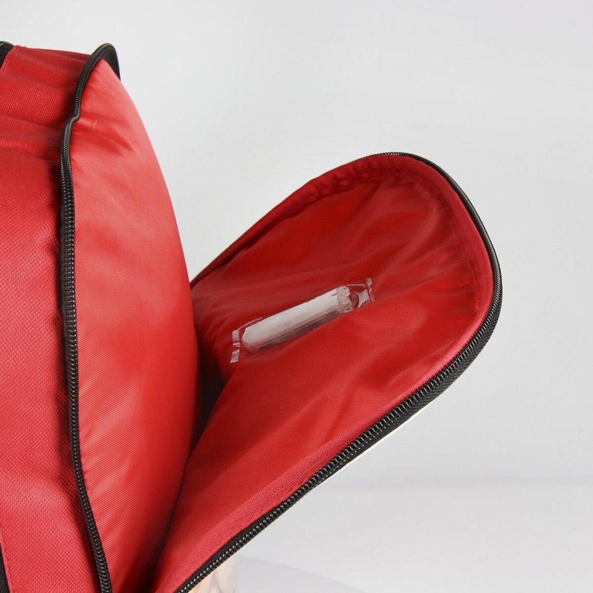 Mochila roja abierta con cremallera negra. Dentro, un paquete de plástico transparente es visible contra el fondo de tela roja. Fondo blanco.