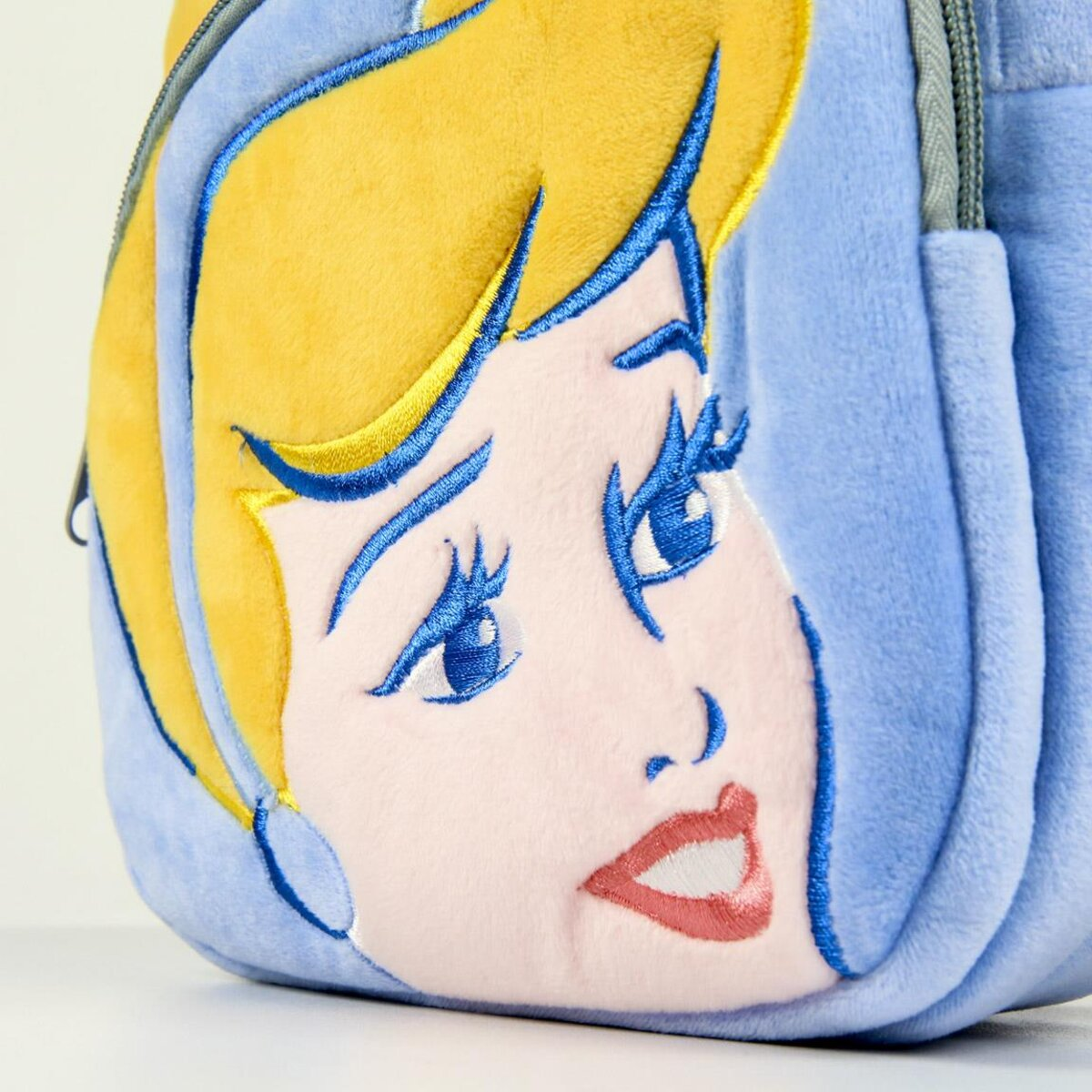 Primer plano de una mochila azul con la cara de Cenicienta. Cabello rubio, ojos azules y boca rosada. La cremallera es visible.