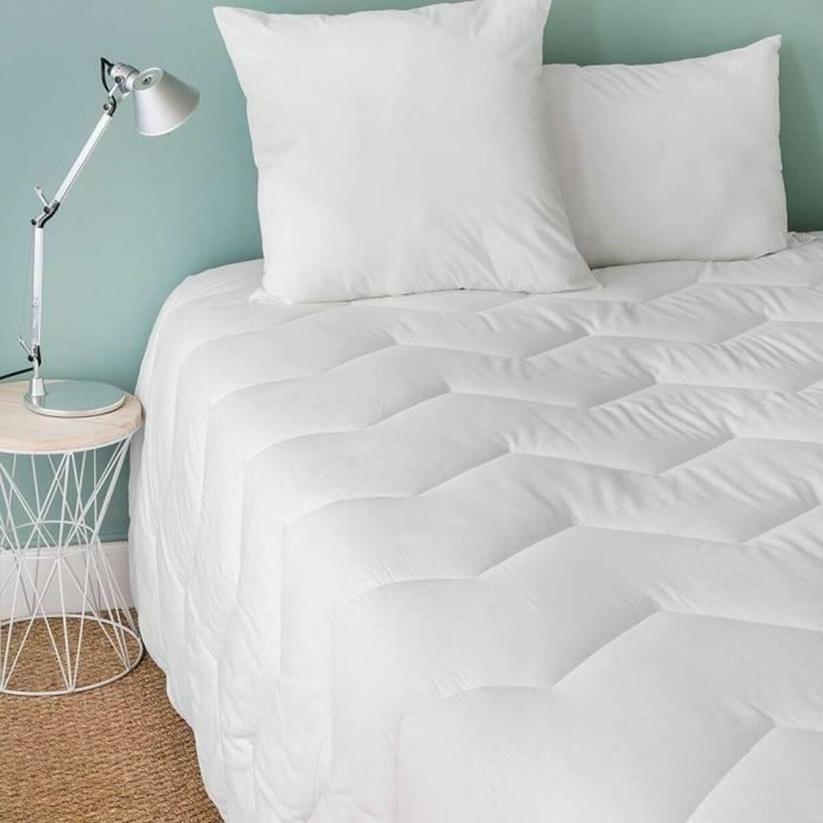 Weißes Bett mit Kissen und einer gemusterten Bettdecke. Ein Beistelltisch und eine Lampe stehen daneben.