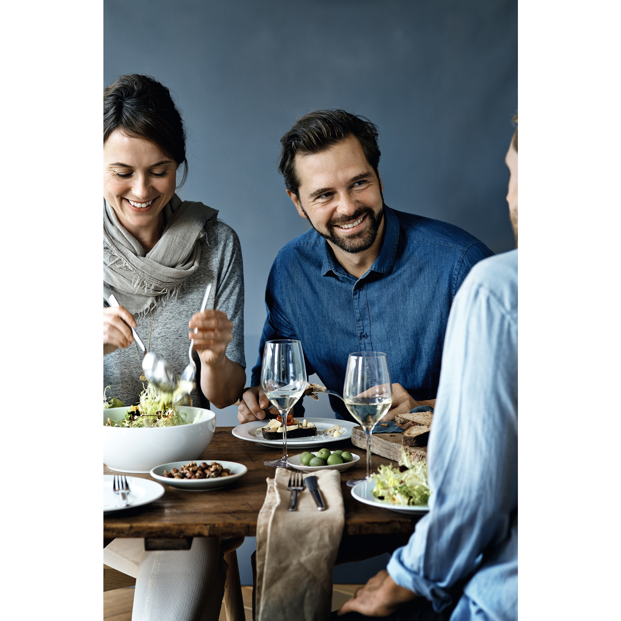 Drei Personen an einem Esstisch, zwei lächeln. Auf dem Tisch stehen Teller, Gläser und Essen.